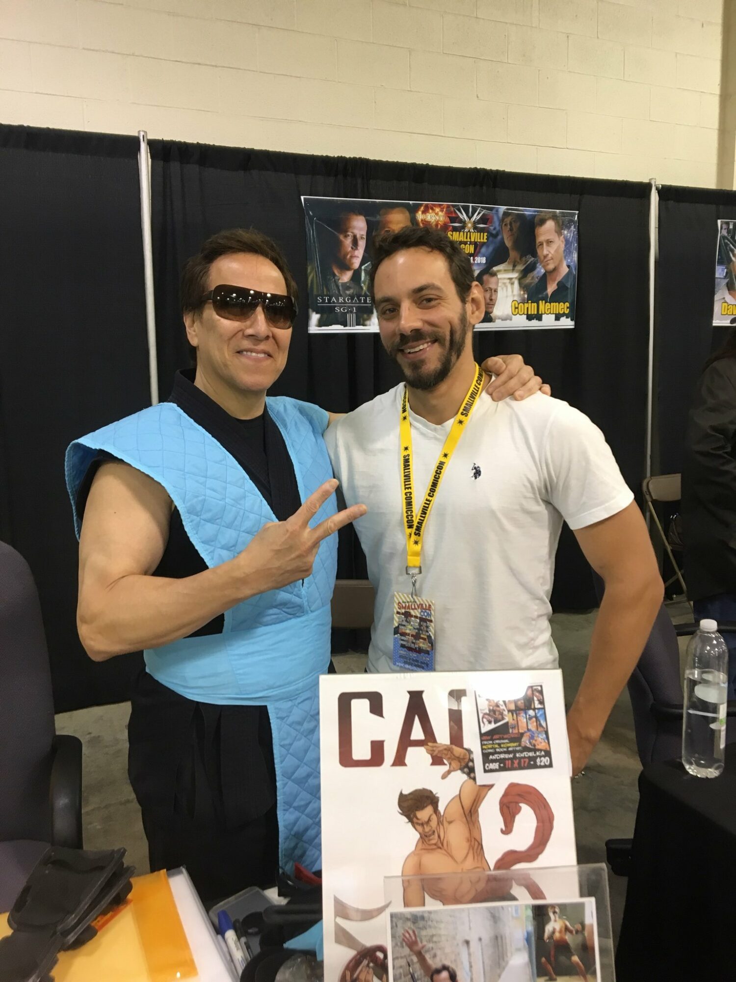 Two men pose together at a convention, one dressed as a character with sunglasses and a blue costume, the other in casual white shirt, both smiling.