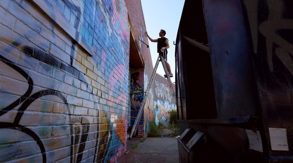Person on a ladder painting graffiti on a brick wall in an alleyway, with a large dark structure nearby.