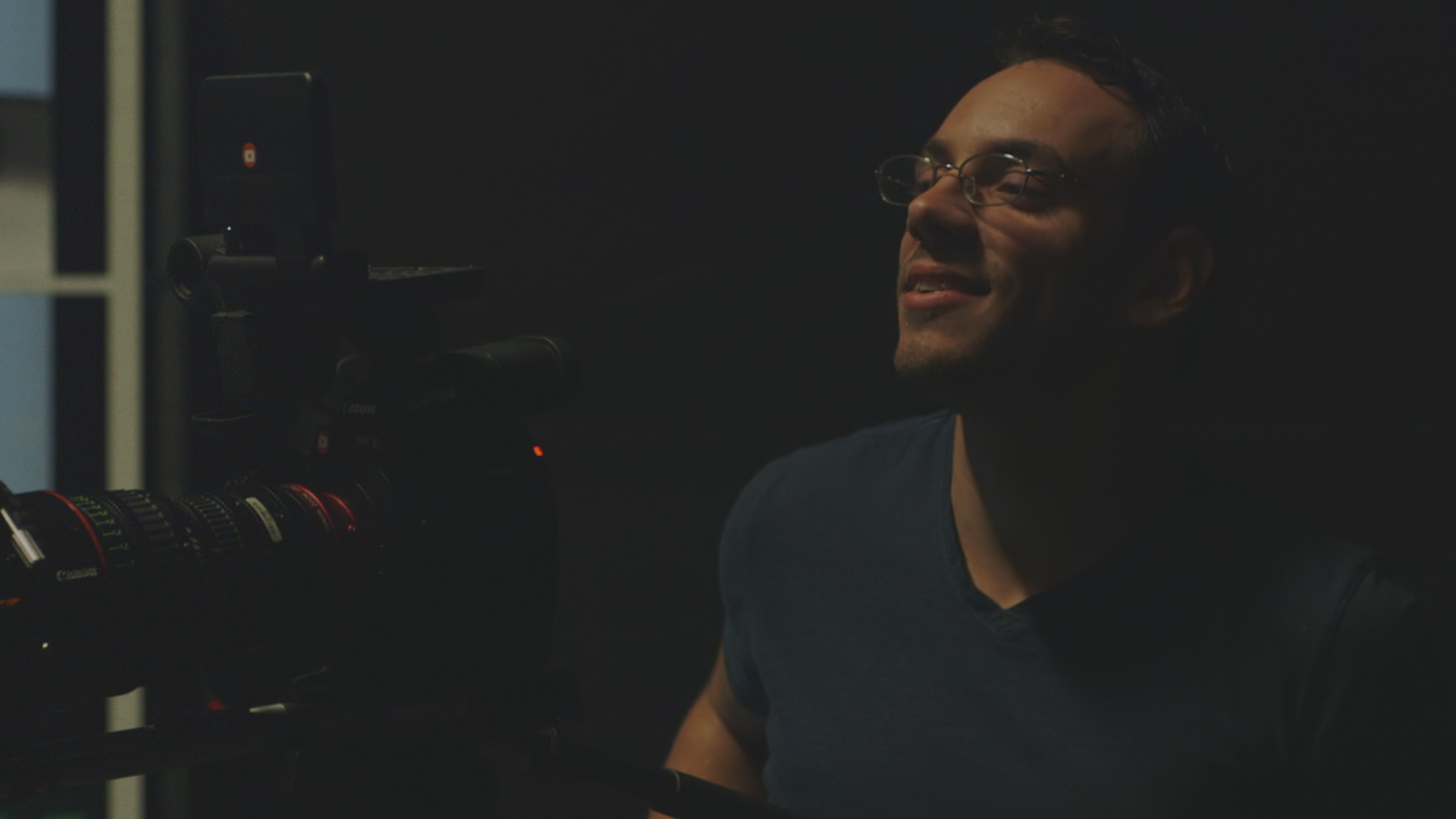 Person with glasses smiling in a dark room, facing a camera or equipment, with a window in the background.