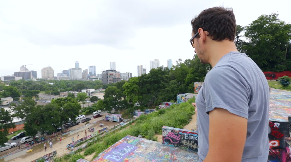 Young man with dark hair and glasses looking at city skyline from a park, overcast sky, trees, and graffiti-covered ledge.