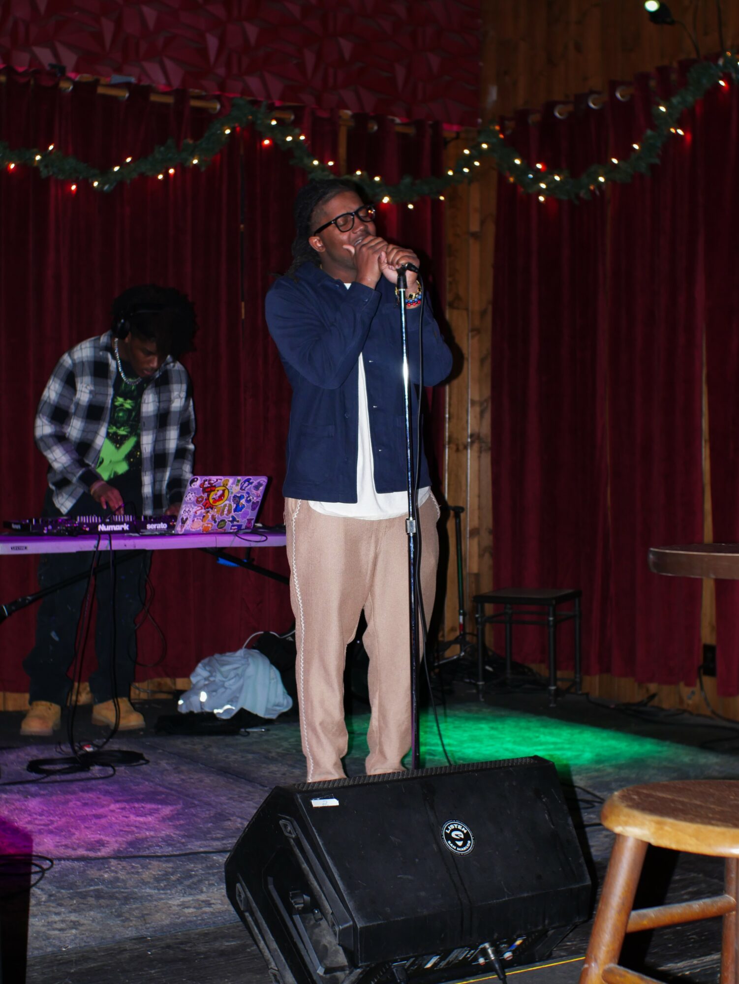 Person singing into a microphone on stage with a DJ in the background, decorated with string lights and red curtains.