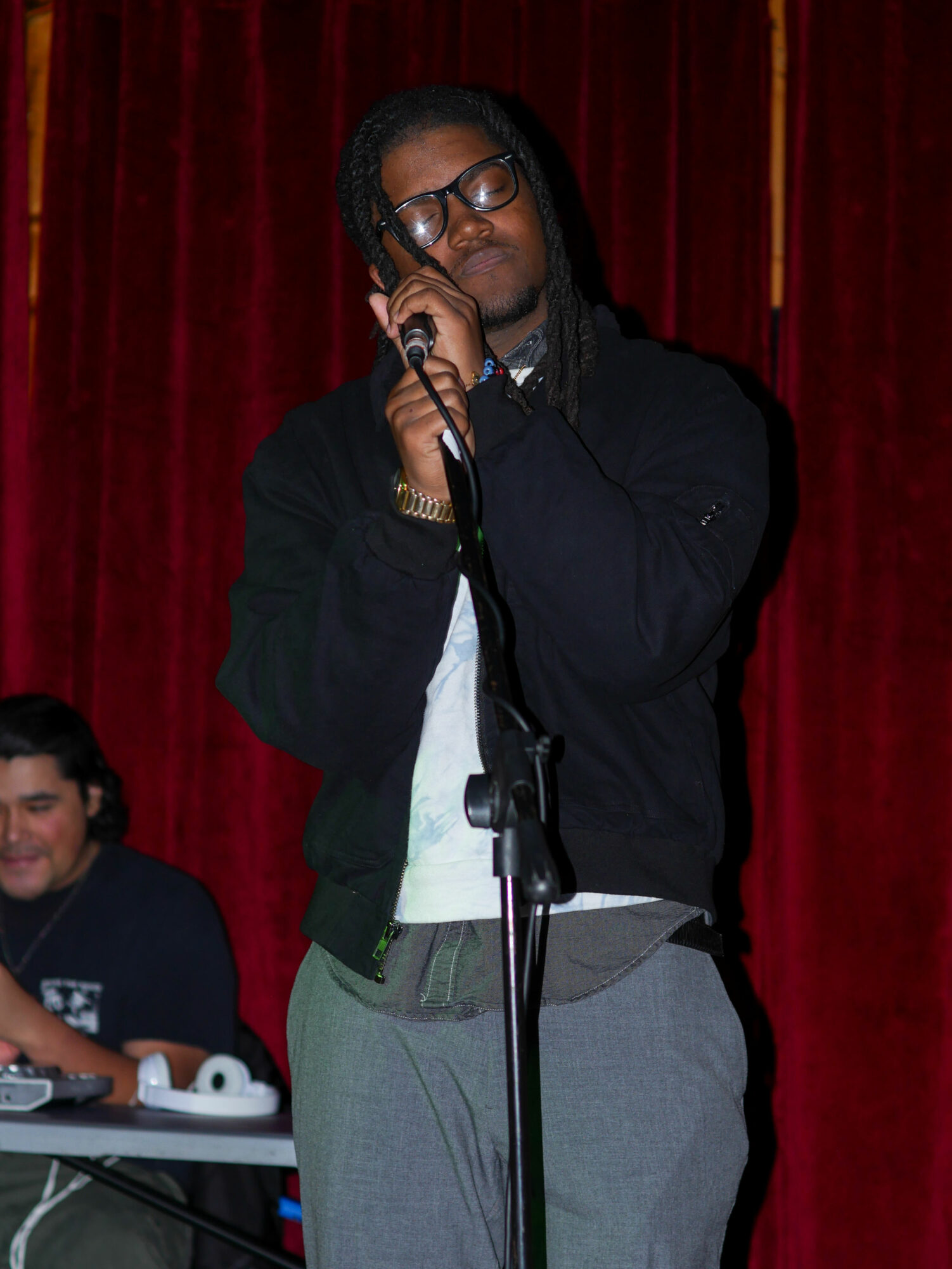 Person with glasses and long hair singing into a microphone on stage with red curtains, another person sitting nearby.