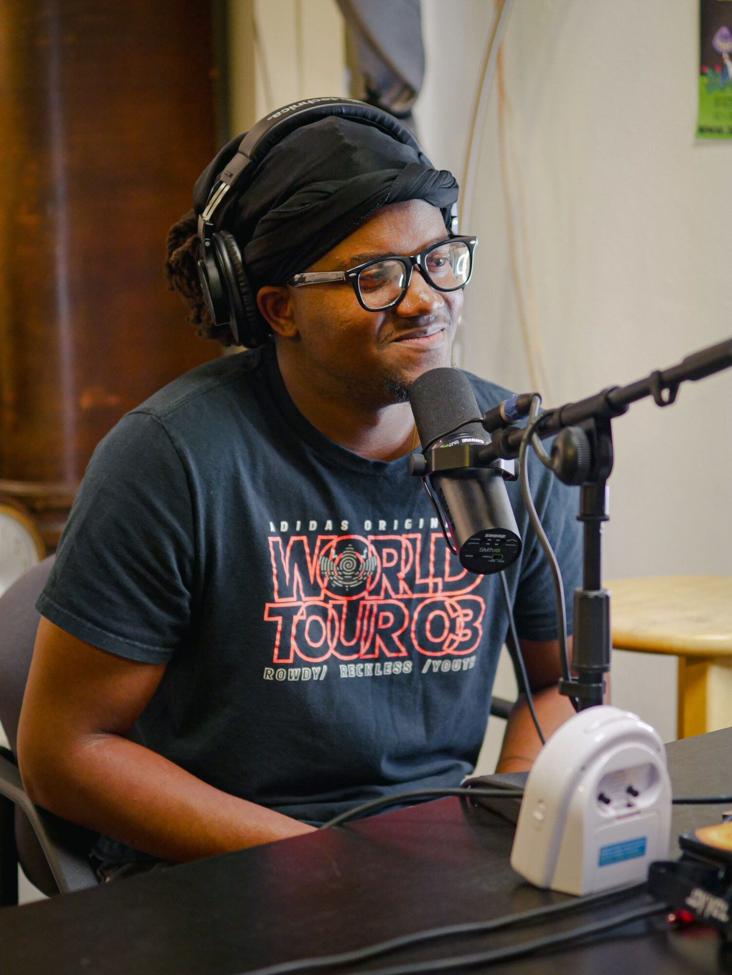 Person wearing glasses and headphones speaking into a microphone in a recording studio.