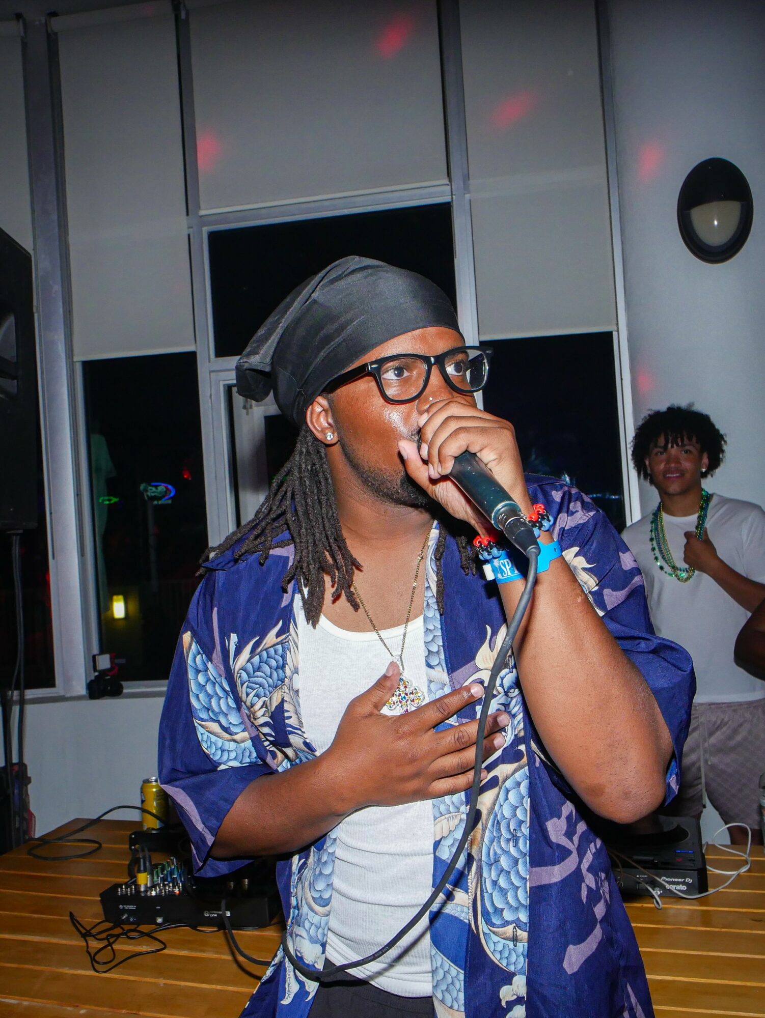 Person with glasses and headscarf singing into a microphone, wearing a blue patterned shirt, in a room with windows and other people.