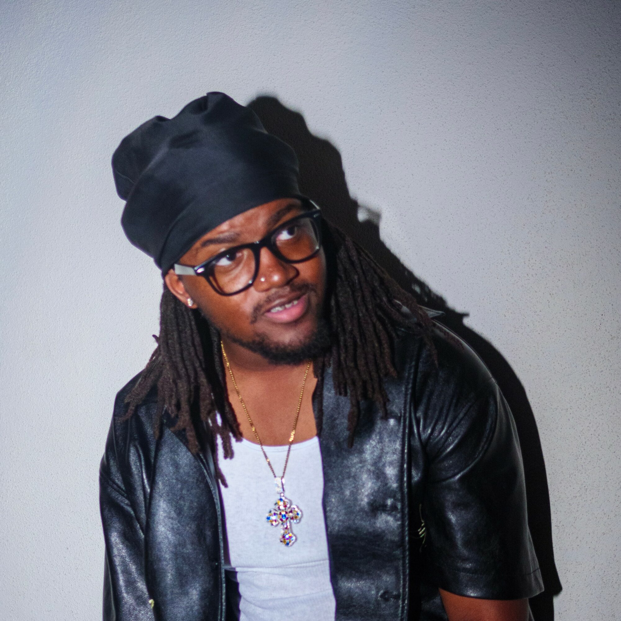 Person with glasses and dreadlocks wearing a black headwrap, leather jacket, and a necklace with a cross pendant, looking to the side.