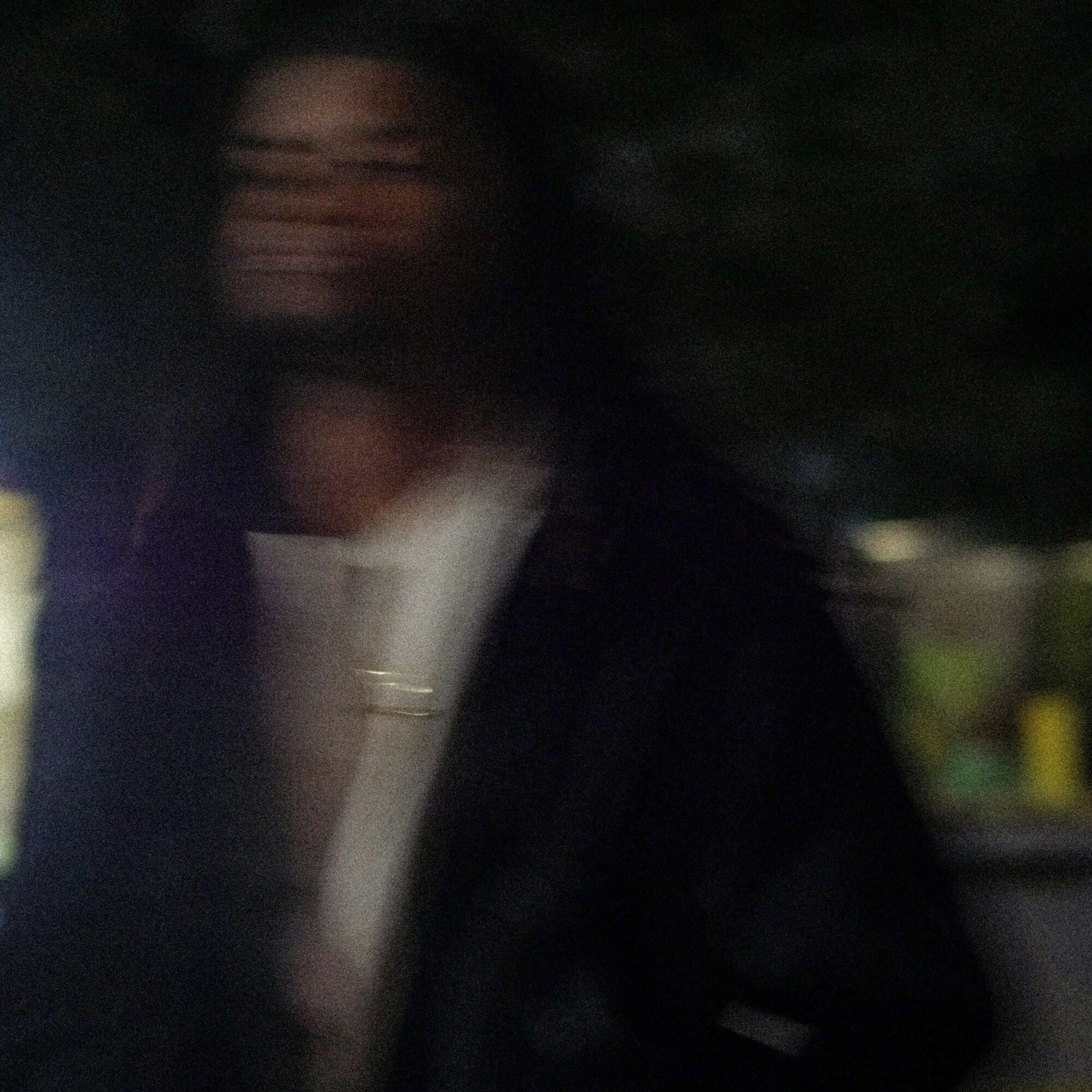 Blurred image of a person with dark hair outdoors at night, wearing a dark jacket and light-colored shirt.