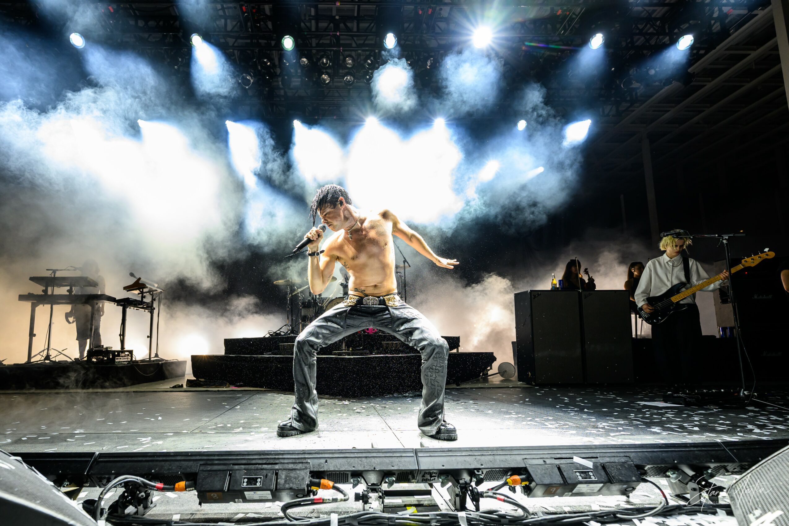 A shirtless performer dances on stage with bright lights and smoke effects, with musicians playing in the background.