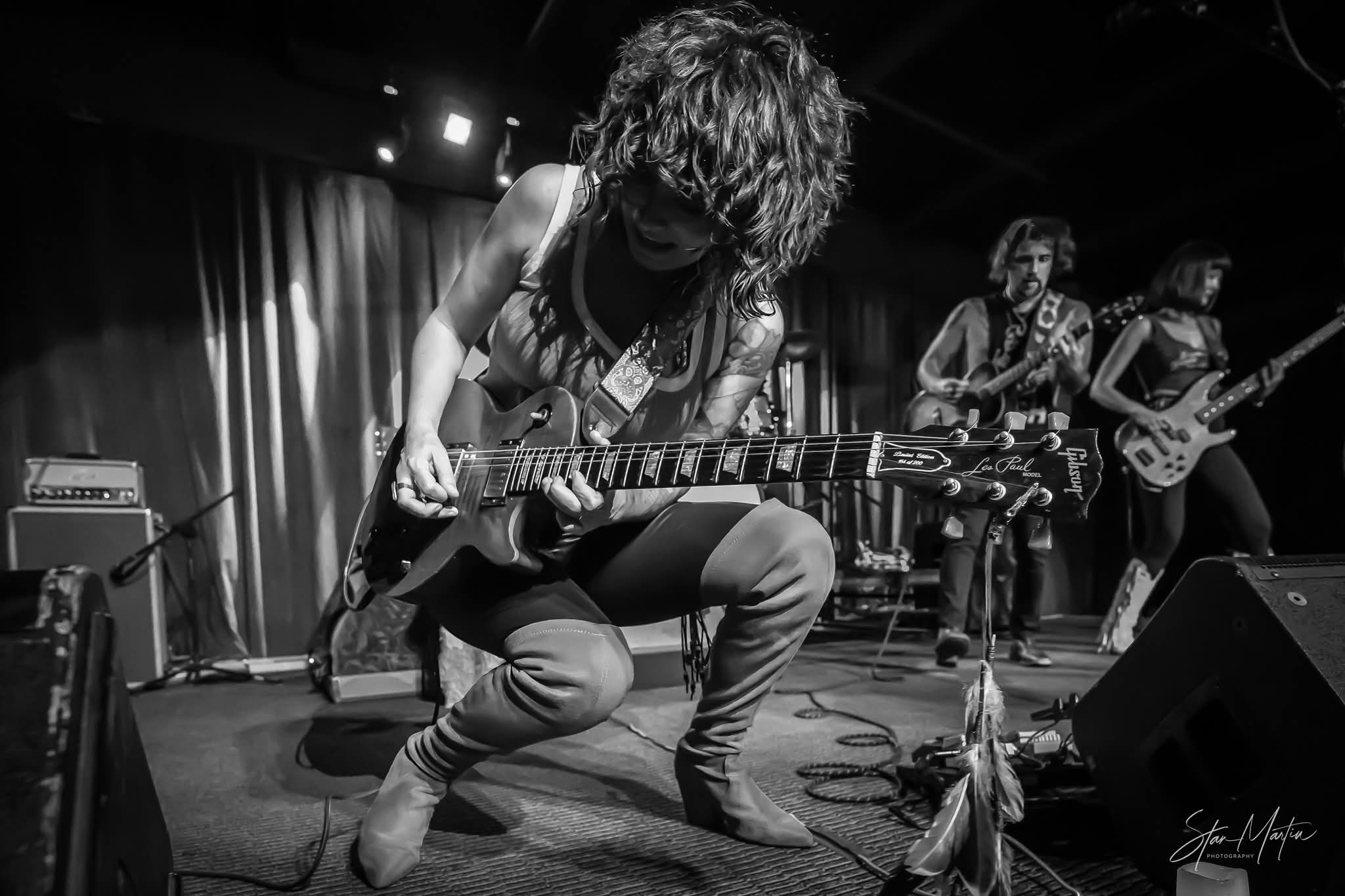 Musician with curly hair playing an electric guitar on stage, with band members in the background, in black and white.