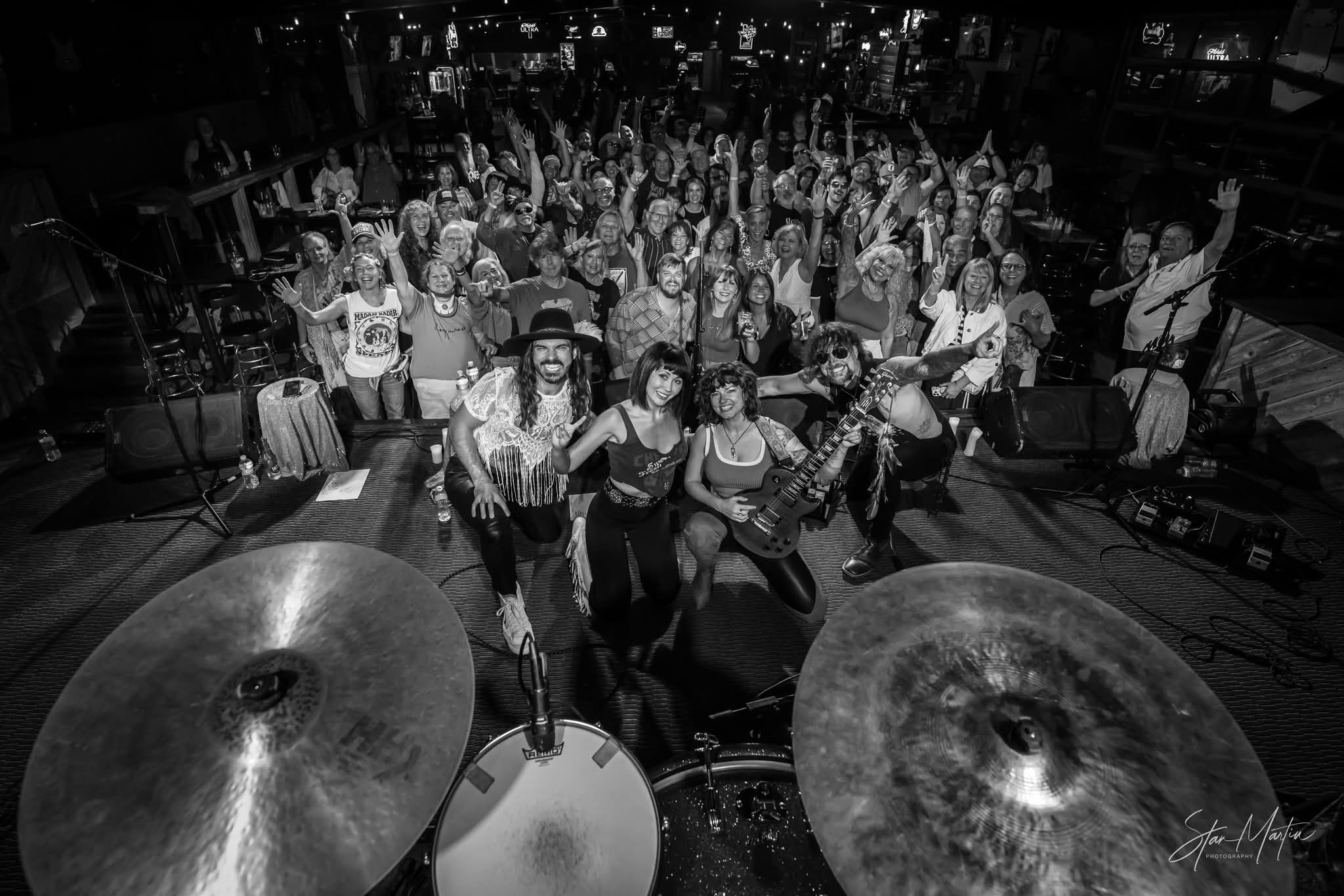 Group of people on stage with musical instruments, audience in background, black and white photo, lively scene.