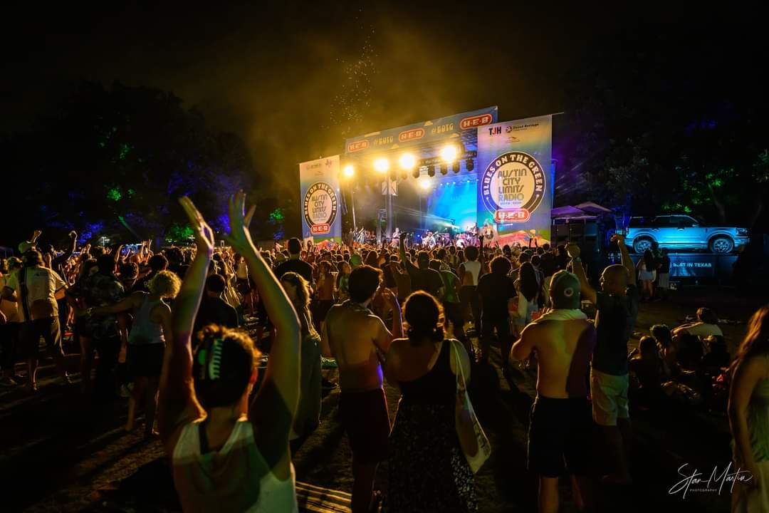 Crowd gathered at an outdoor concert stage with bright lights and banners, night scene with trees in background.