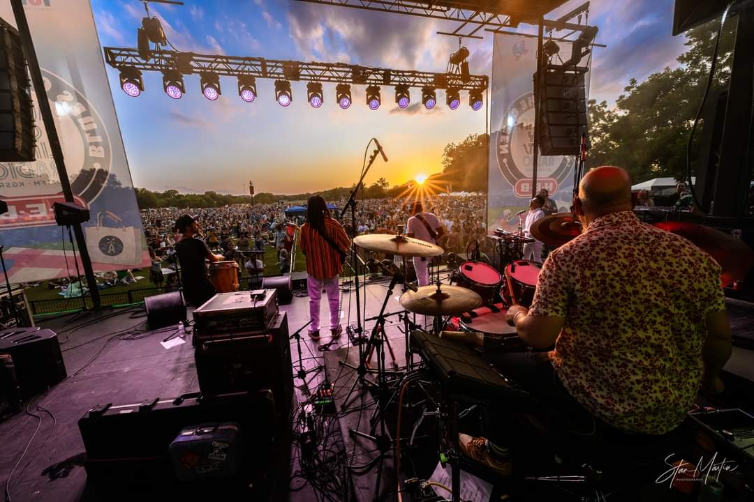 Musicians perform on stage at sunset with a large crowd in the background, stage lights overhead, and trees on the side.