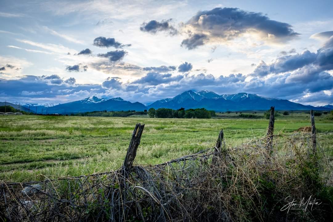 Open field with a fence, mountains in the background, and a cloudy sky.