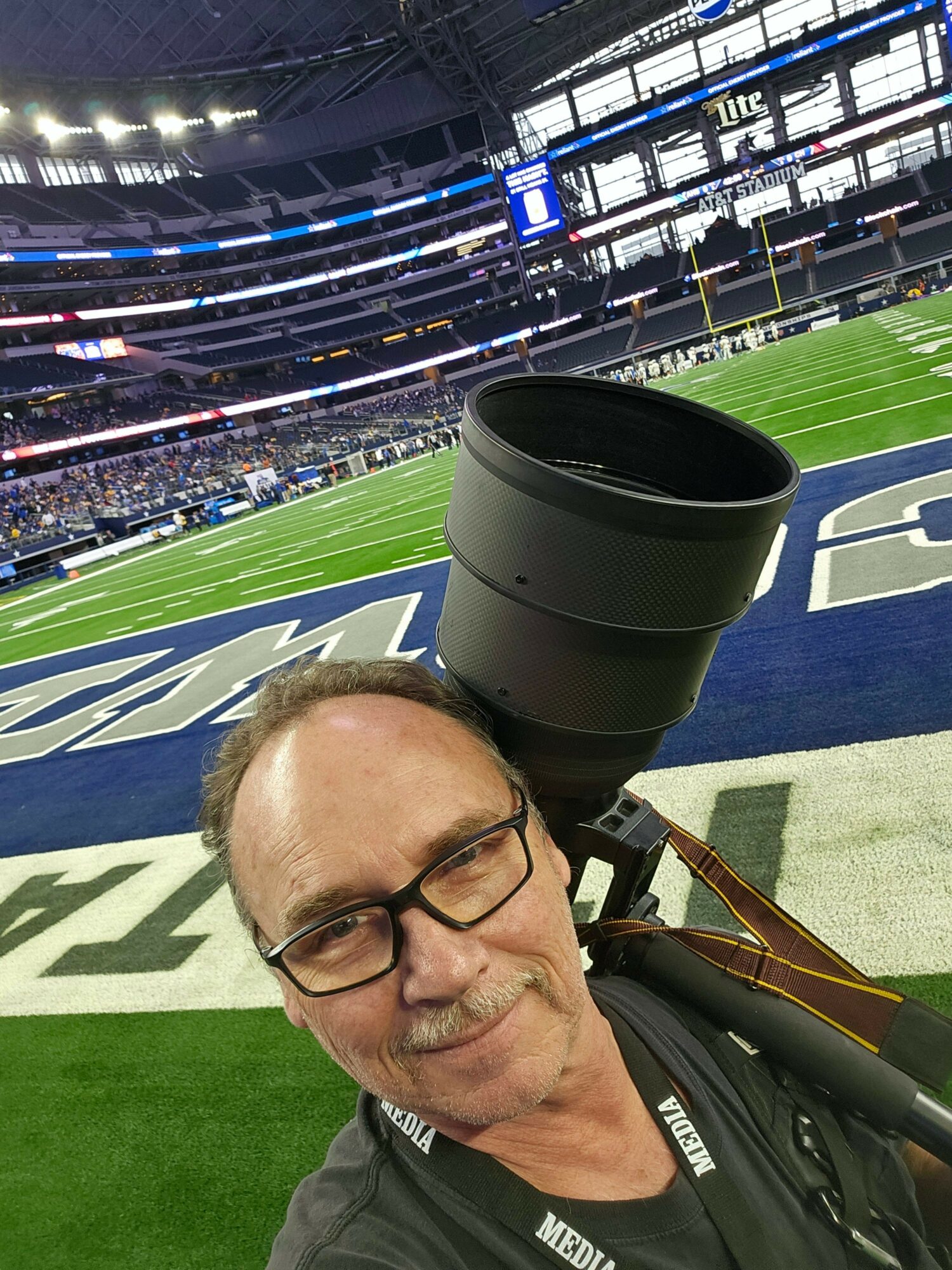 Photographer with glasses and media badge at stadium with field and seating visible