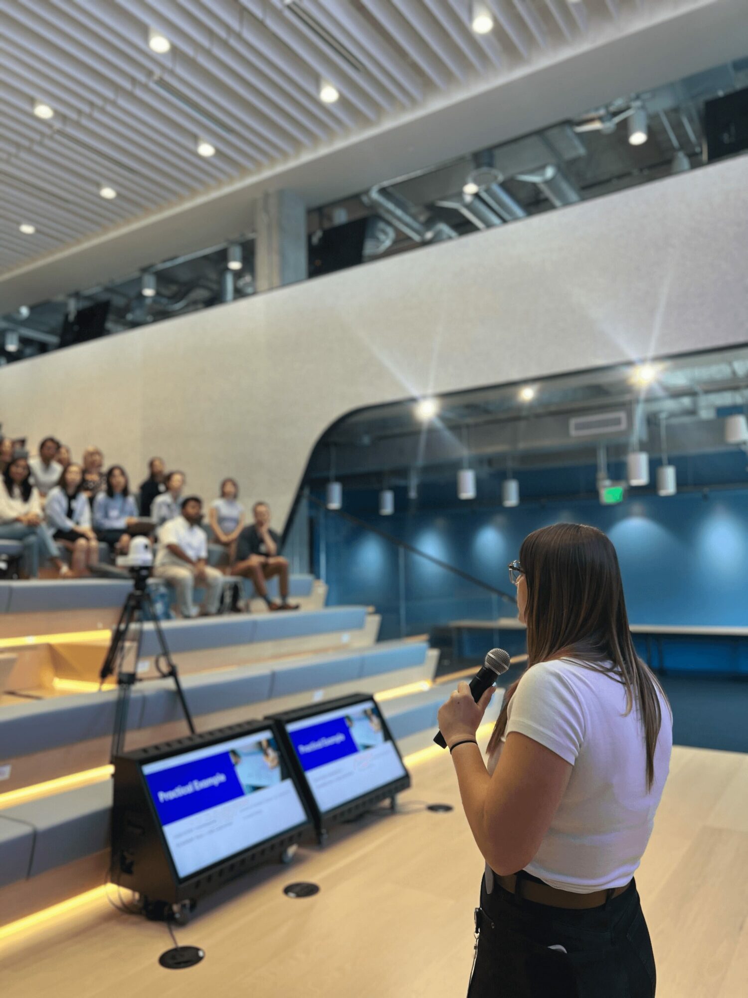 Woman speaking into microphone in front of audience in modern auditorium with tiered seating and blue background.