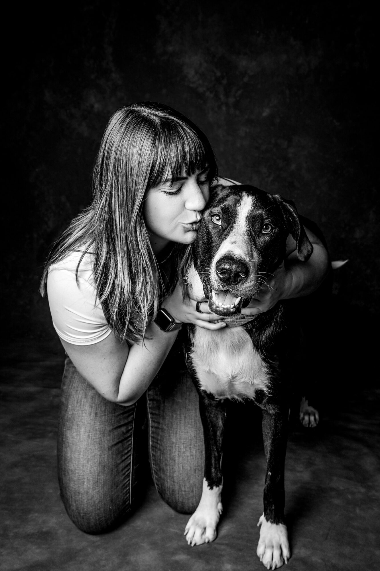 Girl with long hair kissing a large dog on the head, both facing forward, black background, black and white photo.