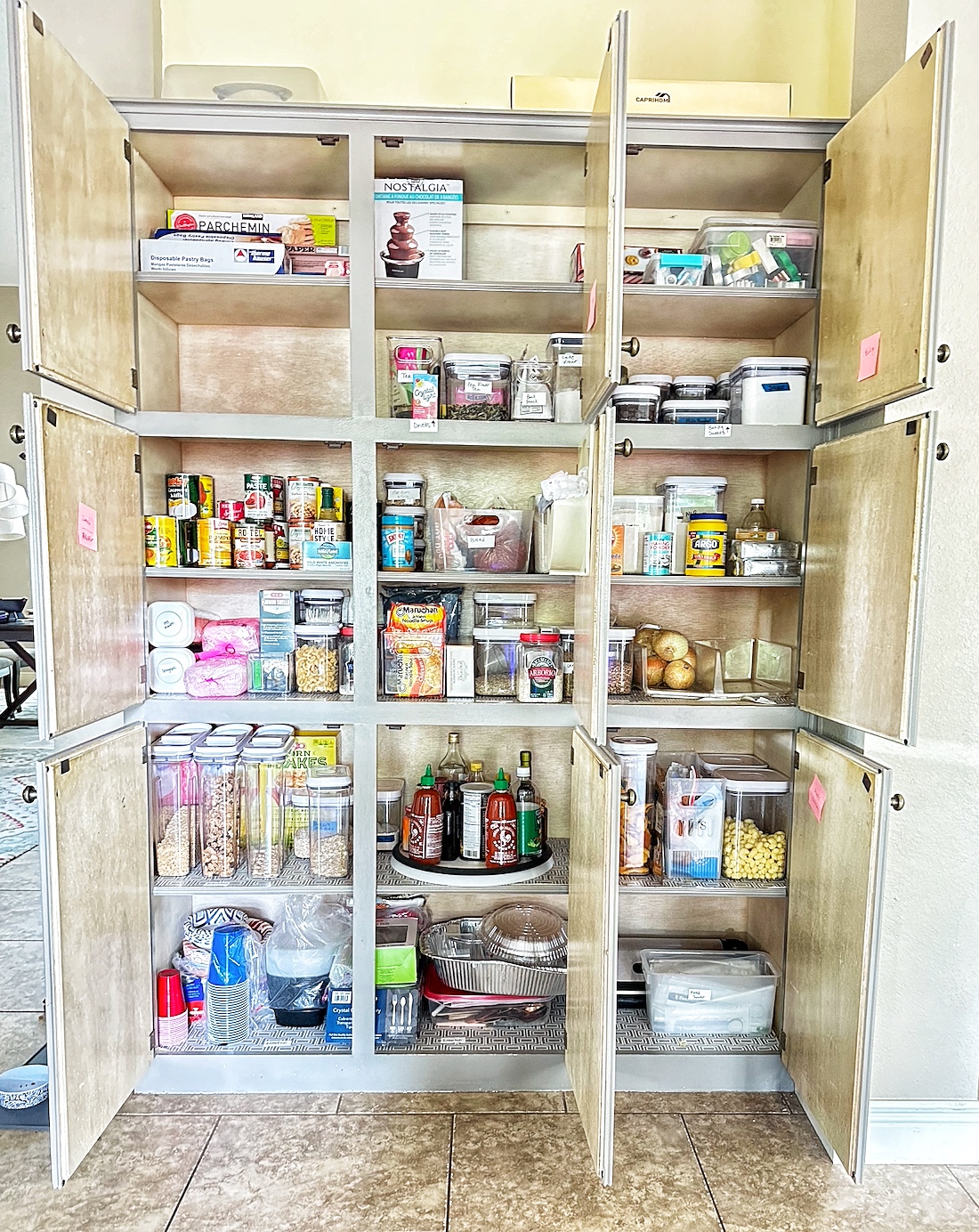Open wooden cabinet with shelves filled with various food and kitchen items, against a tiled floor and light-colored wall.