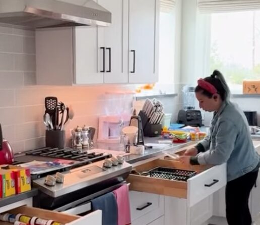 Woman in a kitchen opening a drawer, with cabinets, stove, and window in the background.