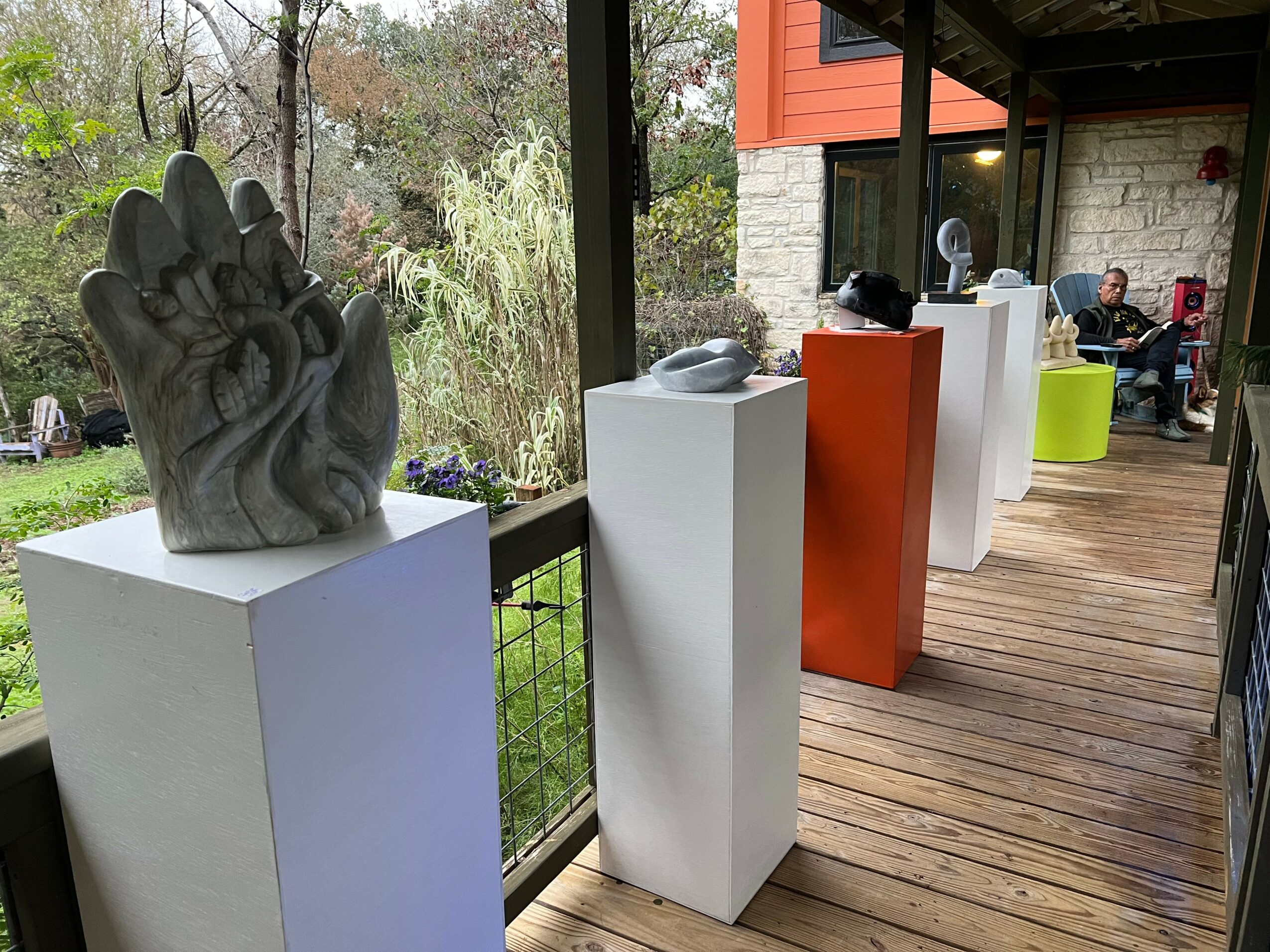 Outdoor porch with sculptures on pedestals, people sitting in the background, wooden floor, and railing, trees visible beyond.