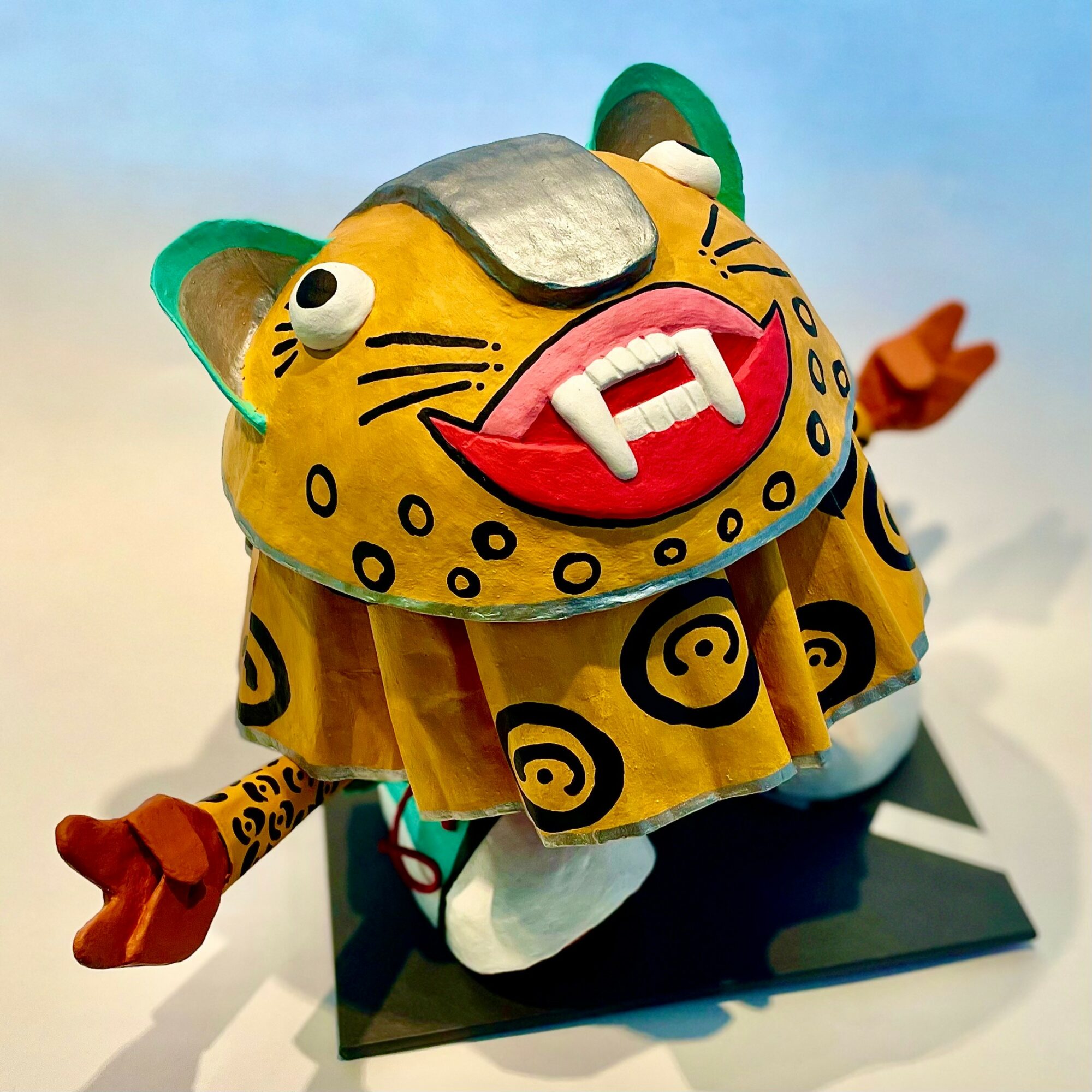 Colorful tiger puppet with glasses, large red mouth, and a yellow patterned body, standing on a black platform against a light background.