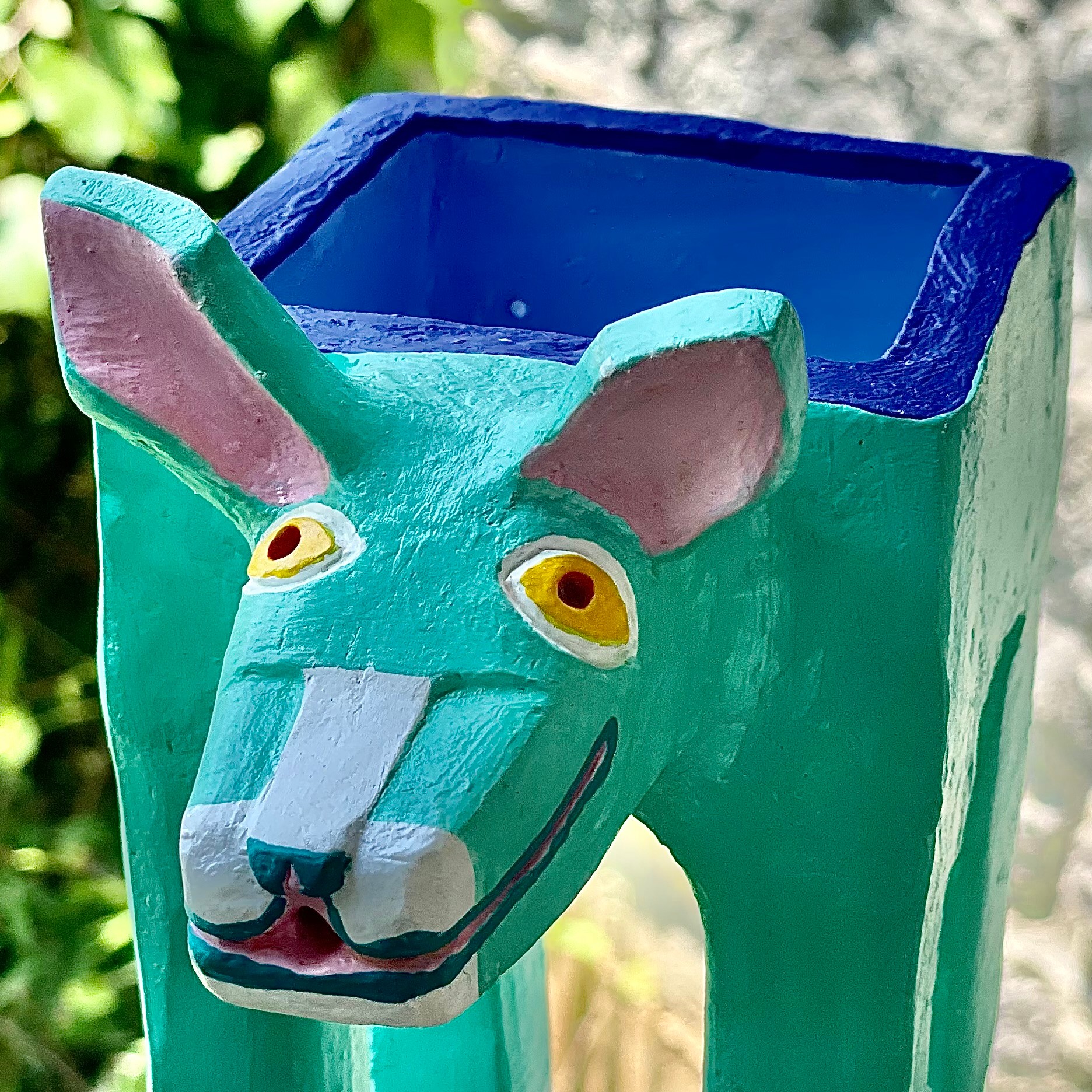 Colorful sculpture of a creature with a lion's face and a rectangular container on its back, outdoors.