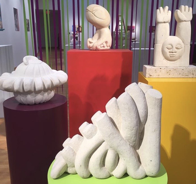 Collection of abstract stone sculptures on colorful pedestals, with a background of vertical purple and green bars.