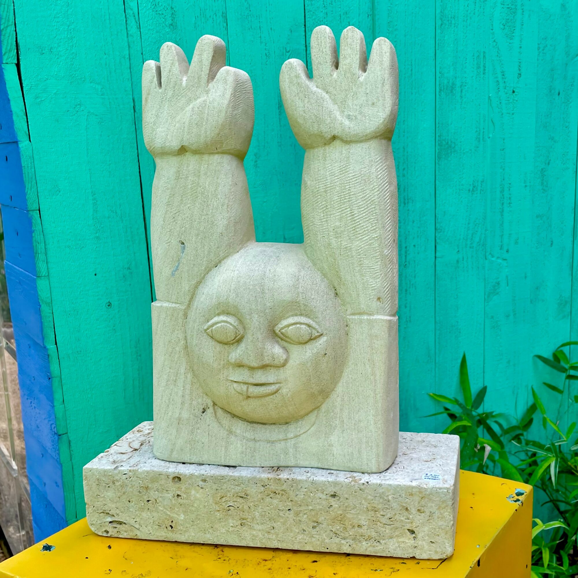 Wooden sculpture of a face with raised hands above a rectangular base, set against a turquoise background with green plants nearby.