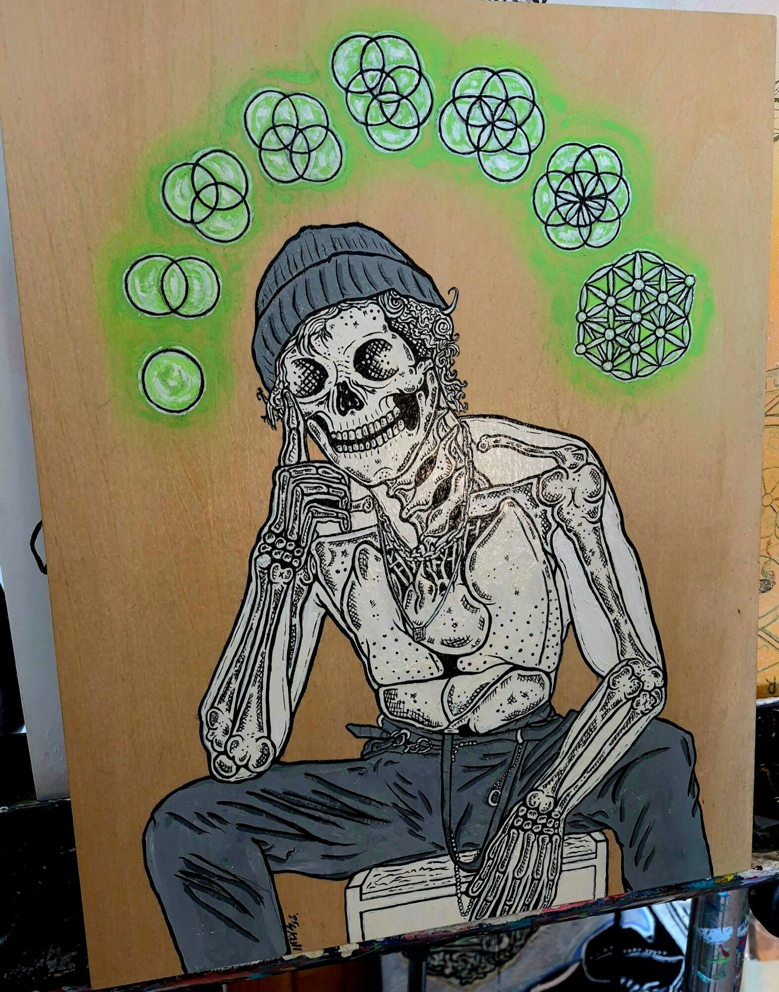 Skeleton wearing a beanie, sitting with one hand on chin, surrounded by geometric patterns in green and black, on a wooden background.