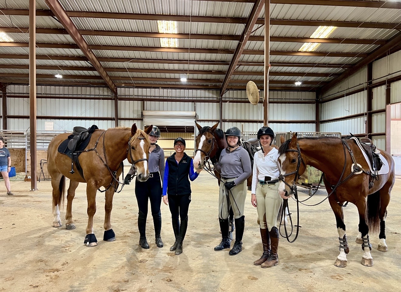 Group of five people standing with three horses inside a riding arena, some holding reins, with a metal roof overhead.