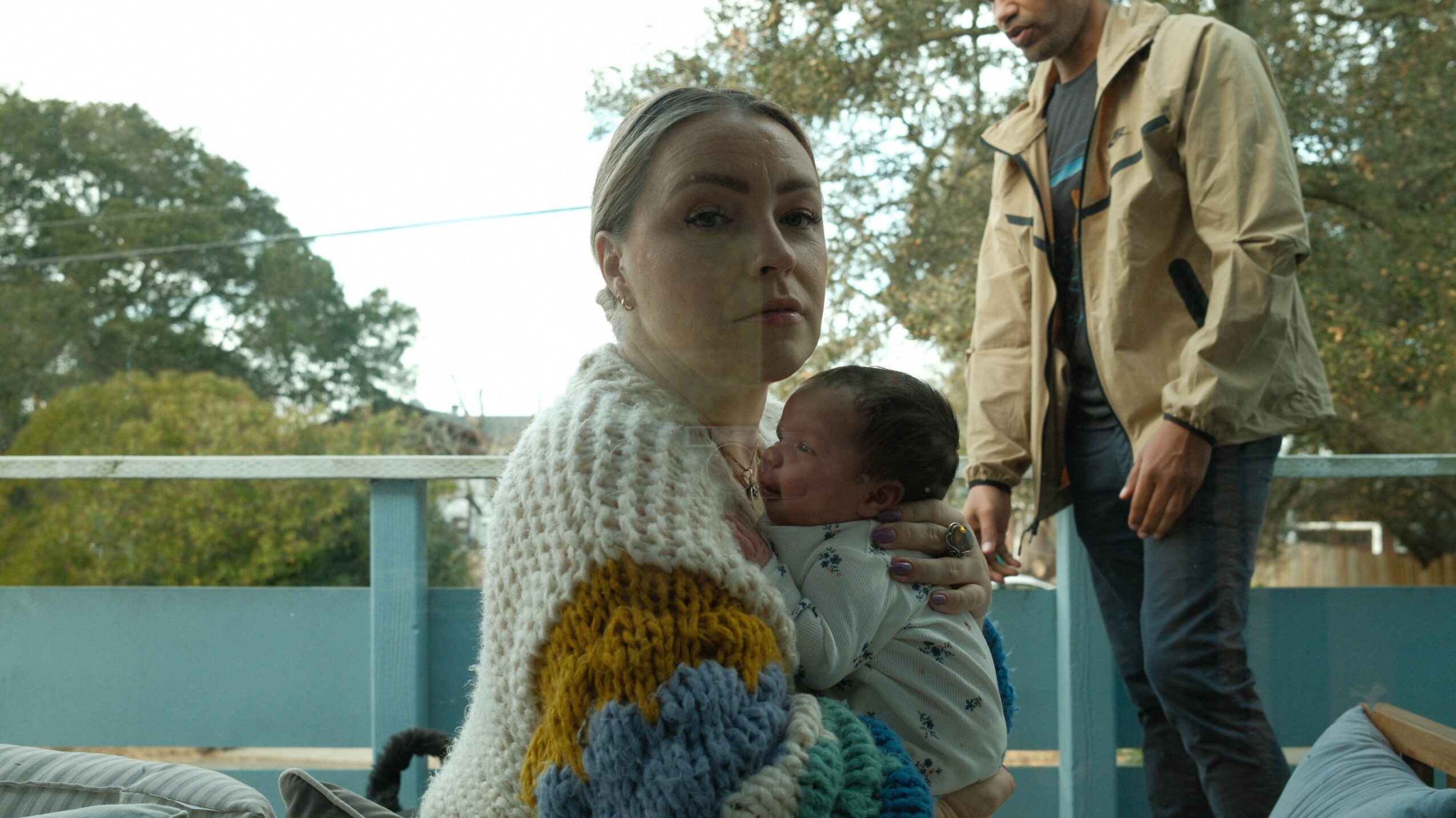 Woman holding a baby, with a man standing nearby outdoors, trees in the background.