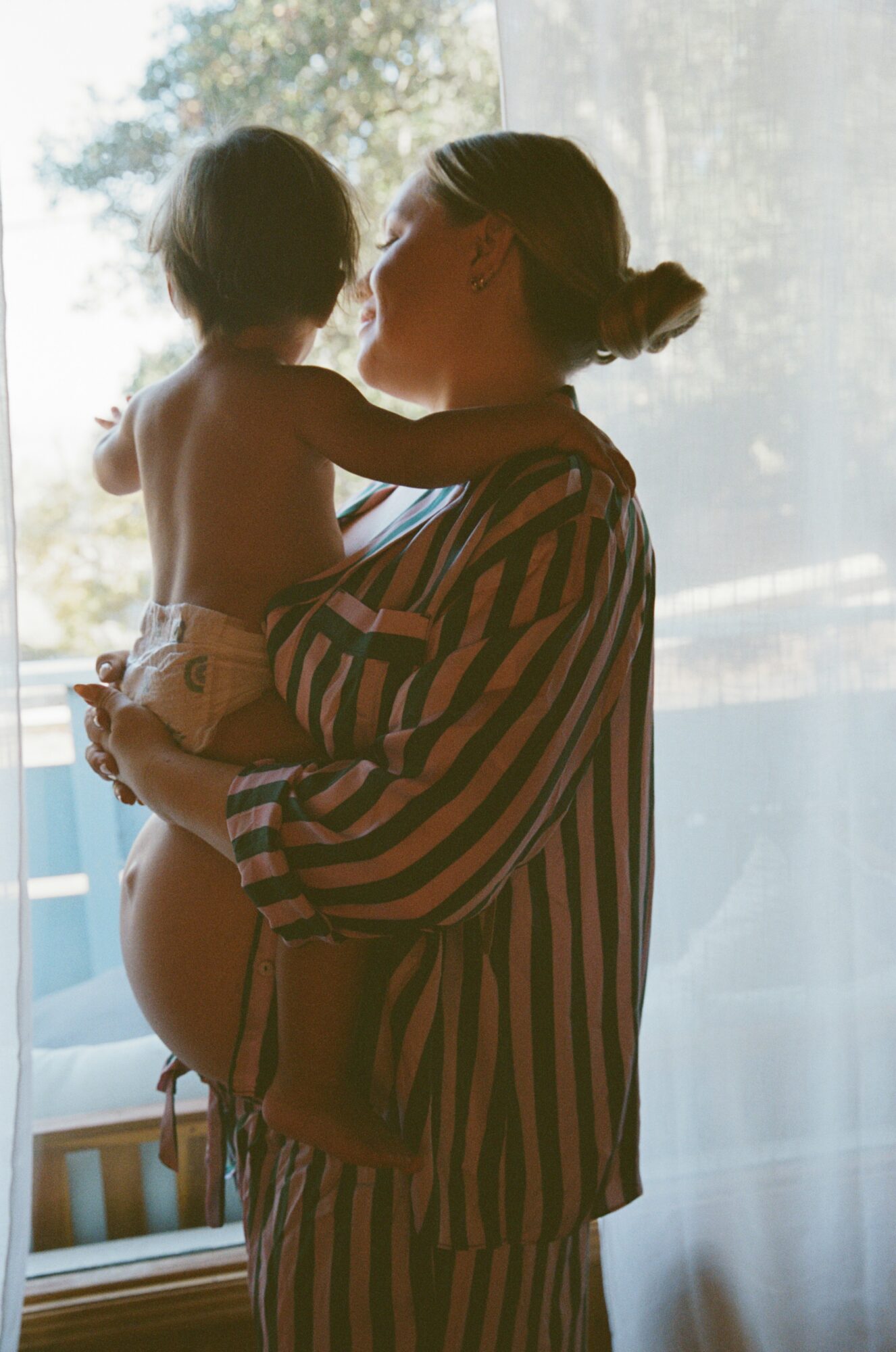Adult holding a child near a window with sunlight, both facing each other, backlit scene, striped shirt visible.