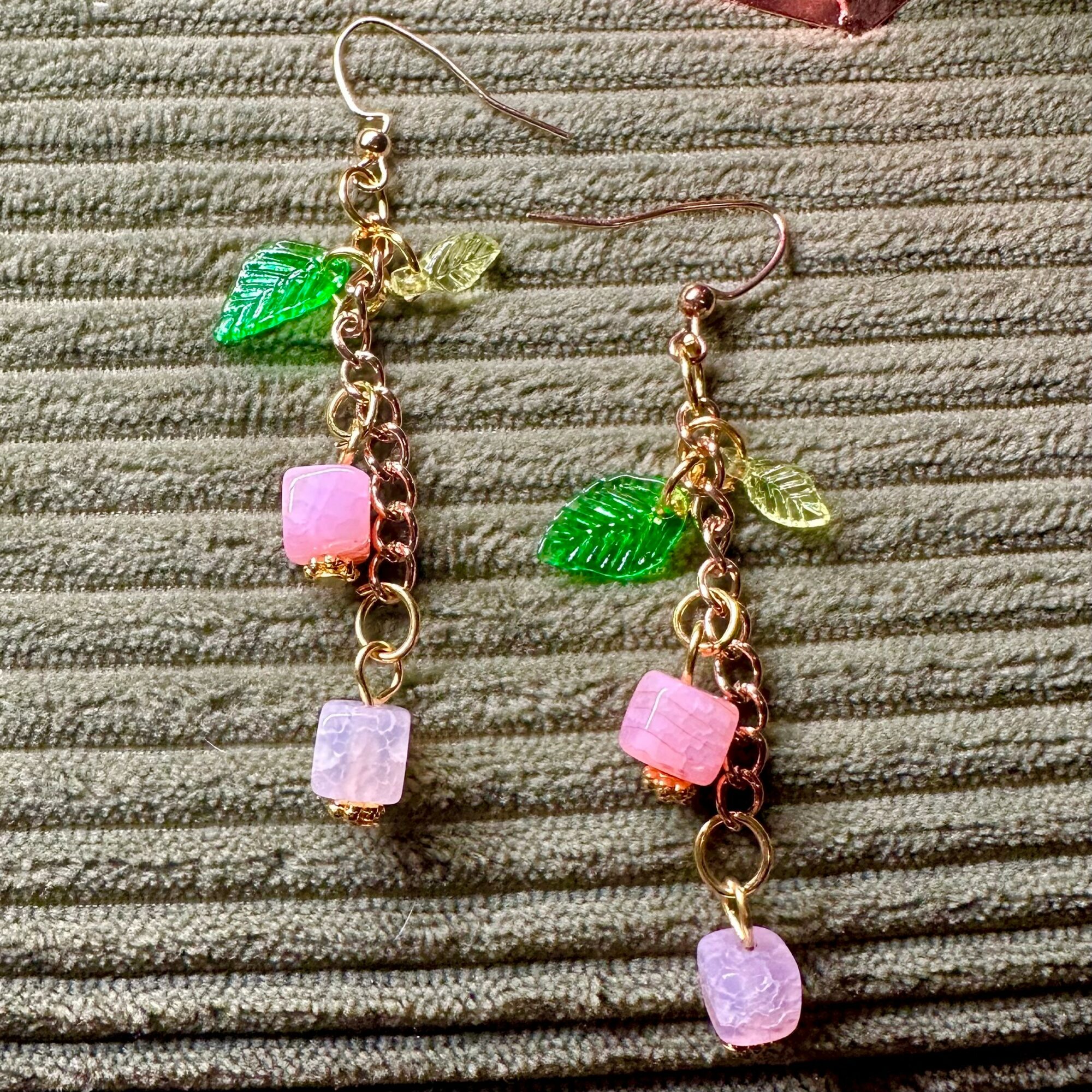 Pair of earrings with pink and purple beads, green leaf charms, and gold chains on textured fabric.