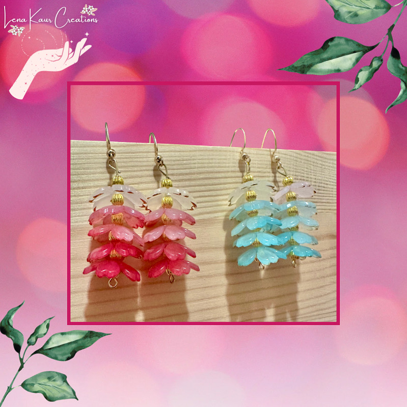 Two pairs of colorful layered earrings, one pink and one blue, hanging on a wooden surface with a pink and purple blurred background.