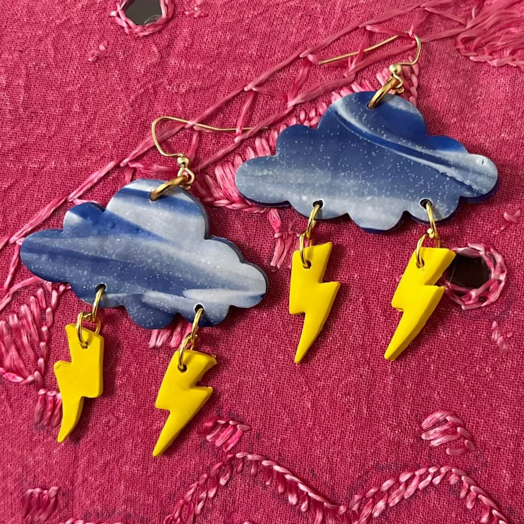 Cloud-shaped earrings with yellow lightning bolt charms on a pink textured background.