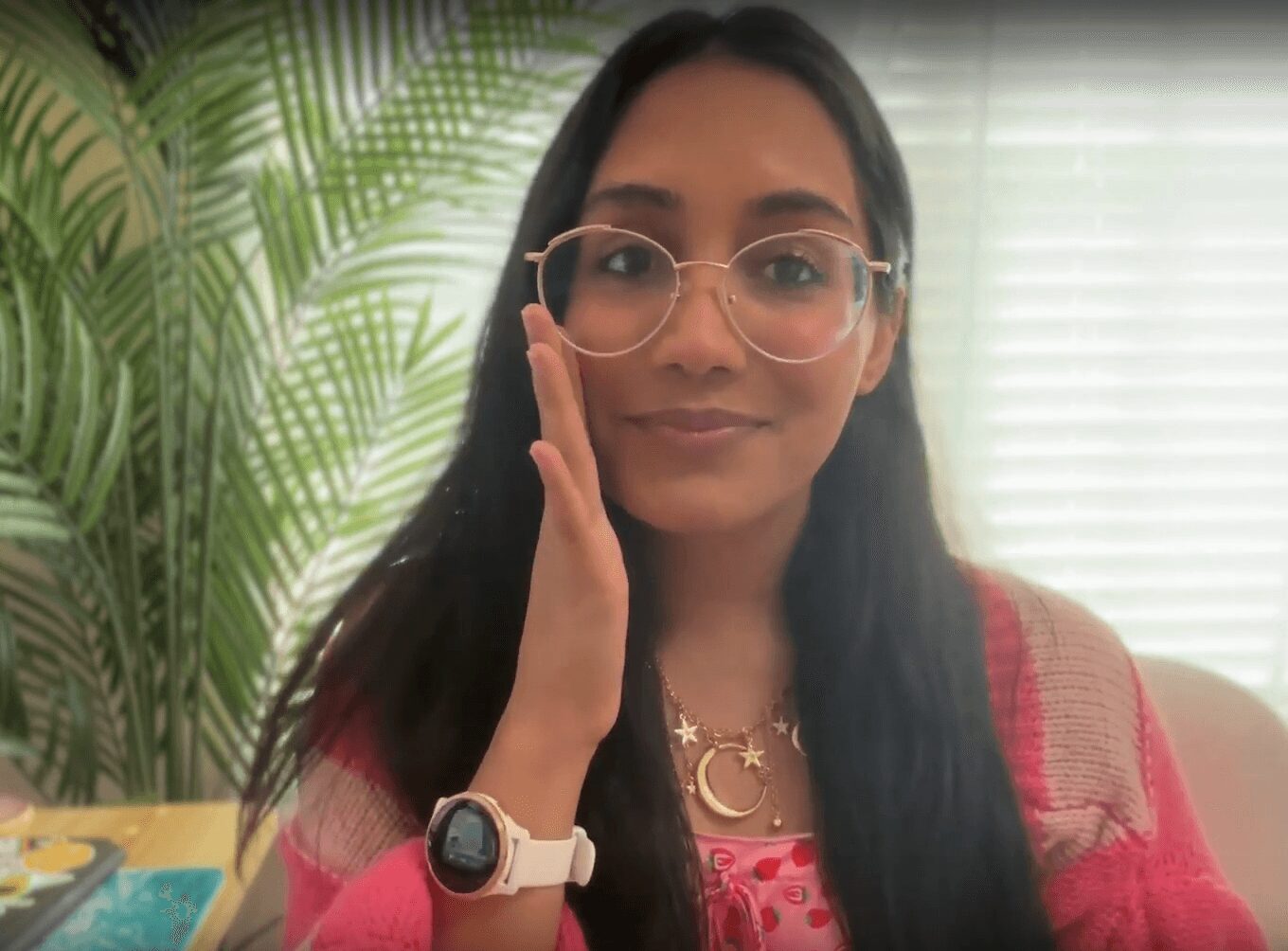Woman with glasses and long dark hair, wearing a pink top and a watch, touching her face, with green plants and window blinds in background.