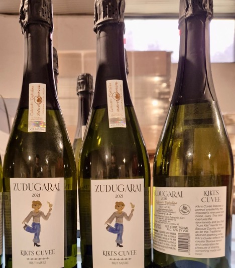 Three bottles of sparkling wine with labels, one showing a cartoon girl dancing, on a kitchen counter near a window.