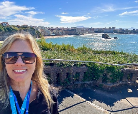 Woman with sunglasses smiling outdoors near water, with a cityscape and blue sky in the background.