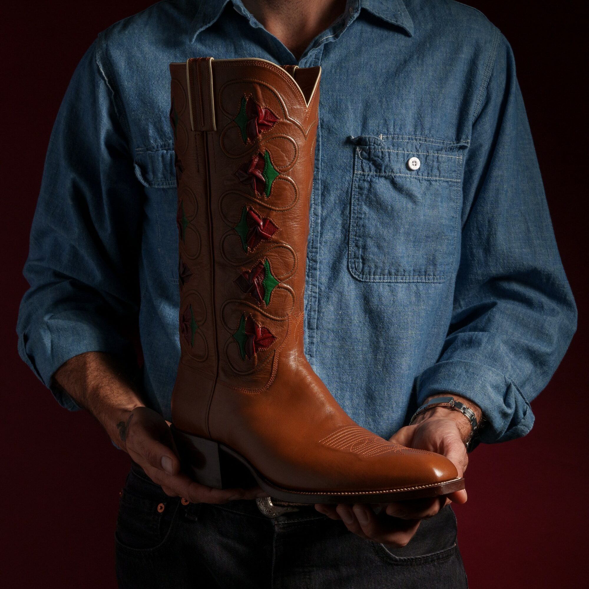 Person in denim shirt holding a tall brown cowboy boot with decorative stitching, against a dark background.