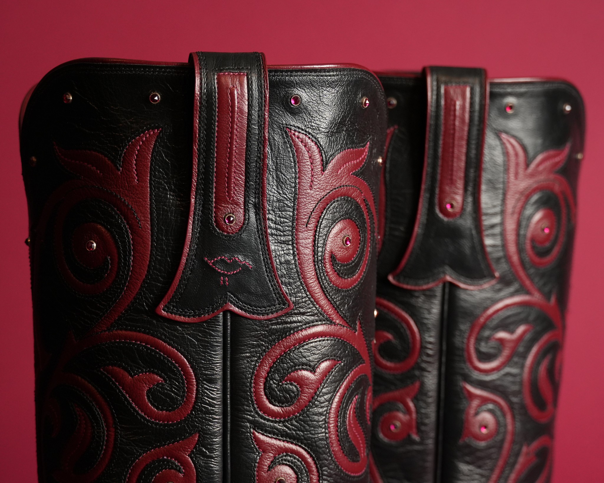 Two black leather boots with red decorative stitching and patterns, standing against a pink background.