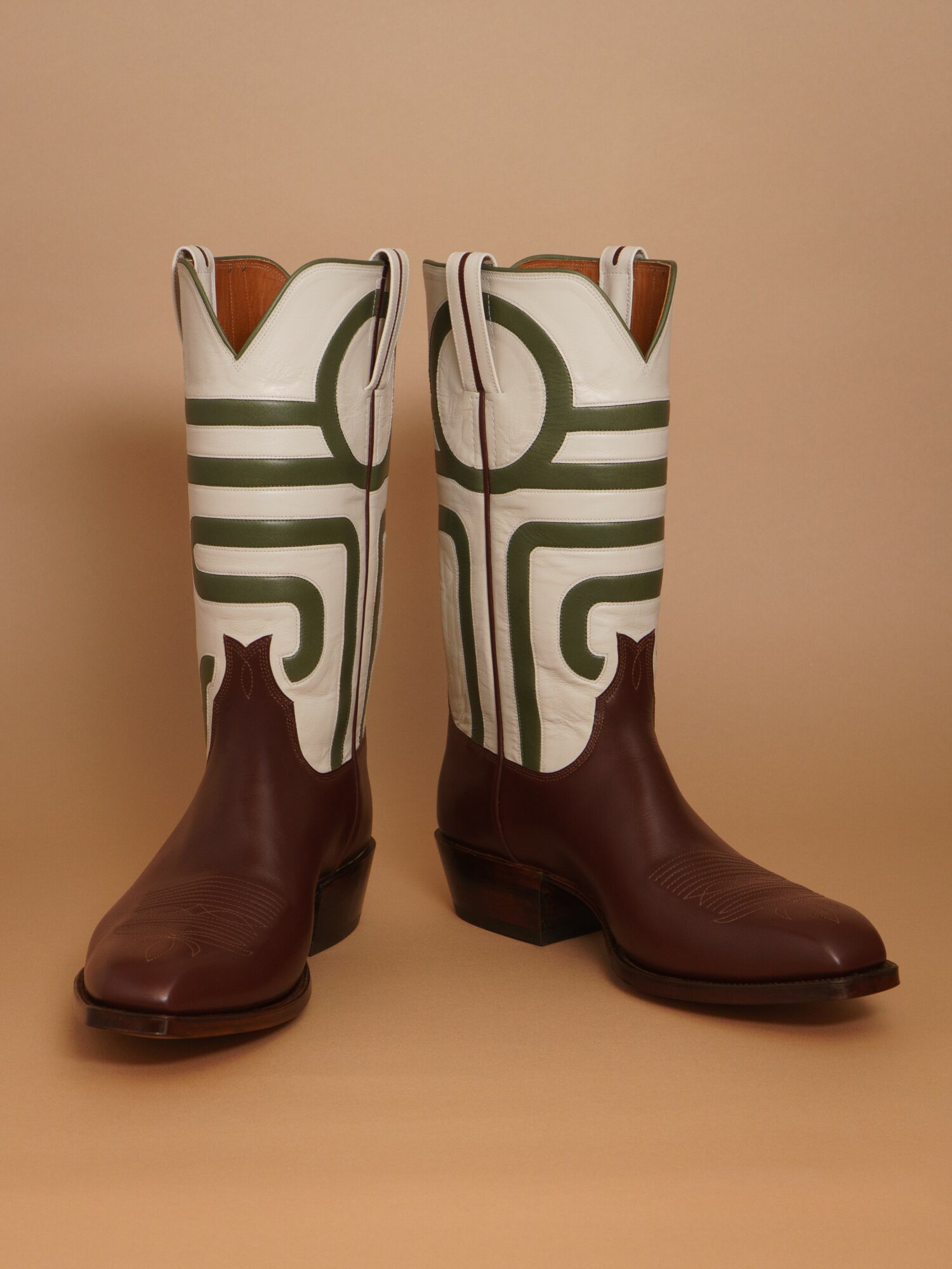Pair of cowboy boots with white, green, and brown design, standing upright against a beige background.