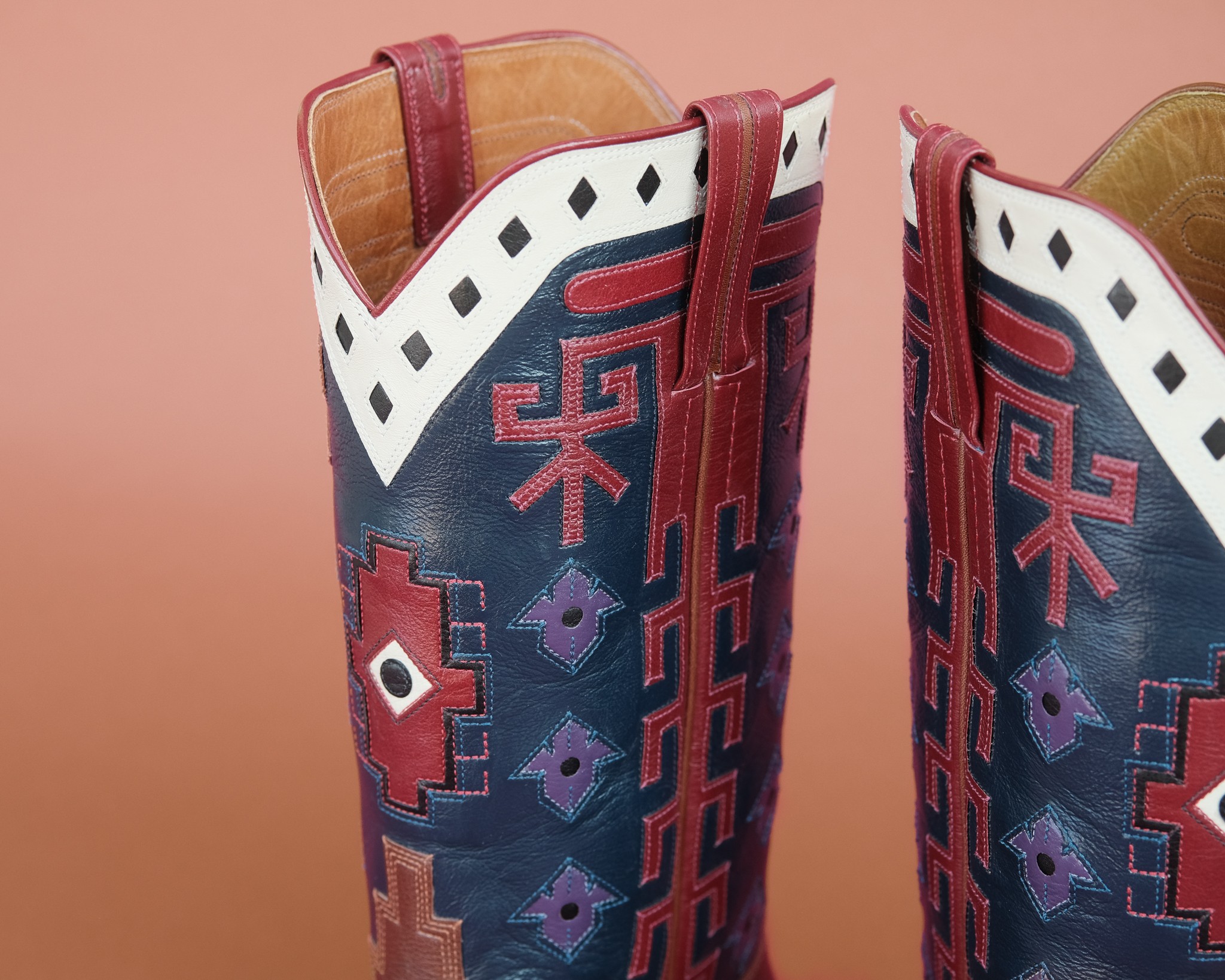 Two cowboy boots with colorful embroidery and a pink background, showing the top part of the boots.