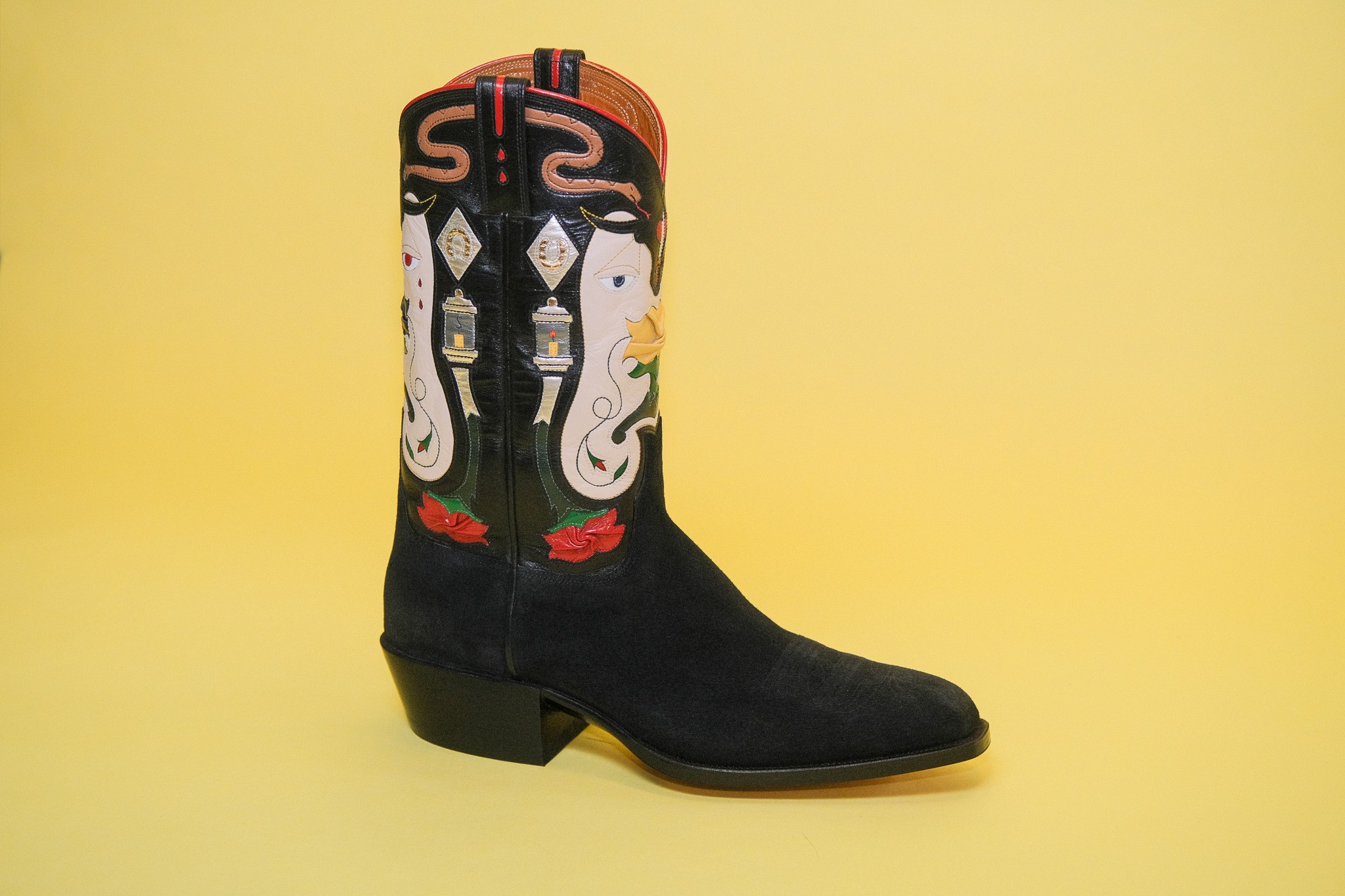 Black cowboy boot with colorful embroidery of faces and patterns, against a yellow background.