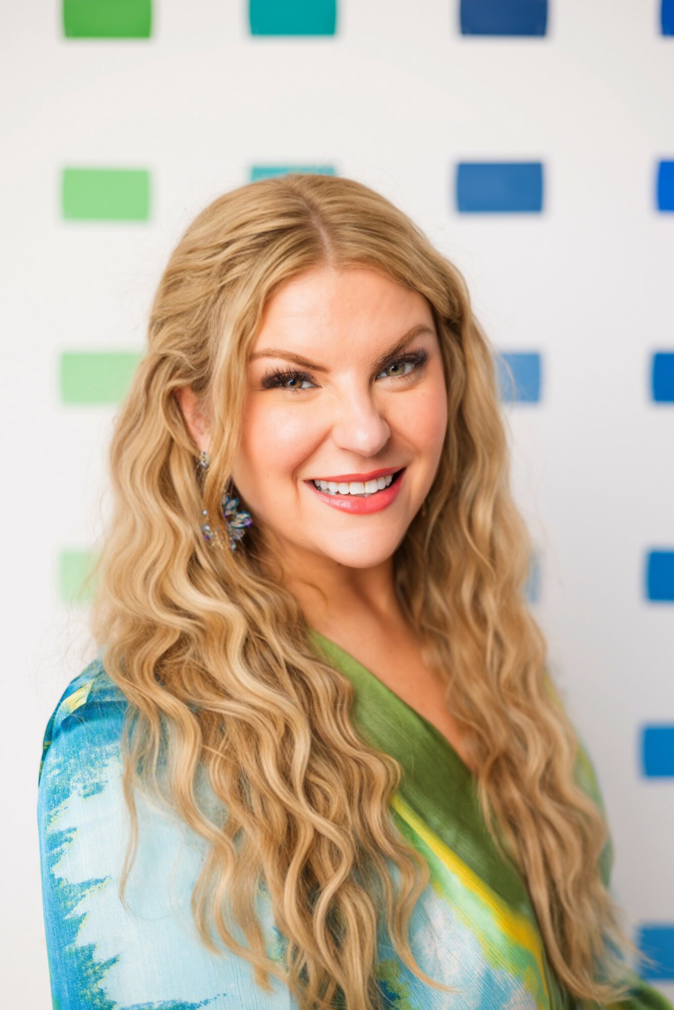Woman with long wavy blonde hair smiling, wearing a colorful top, background with blue and green blocks.