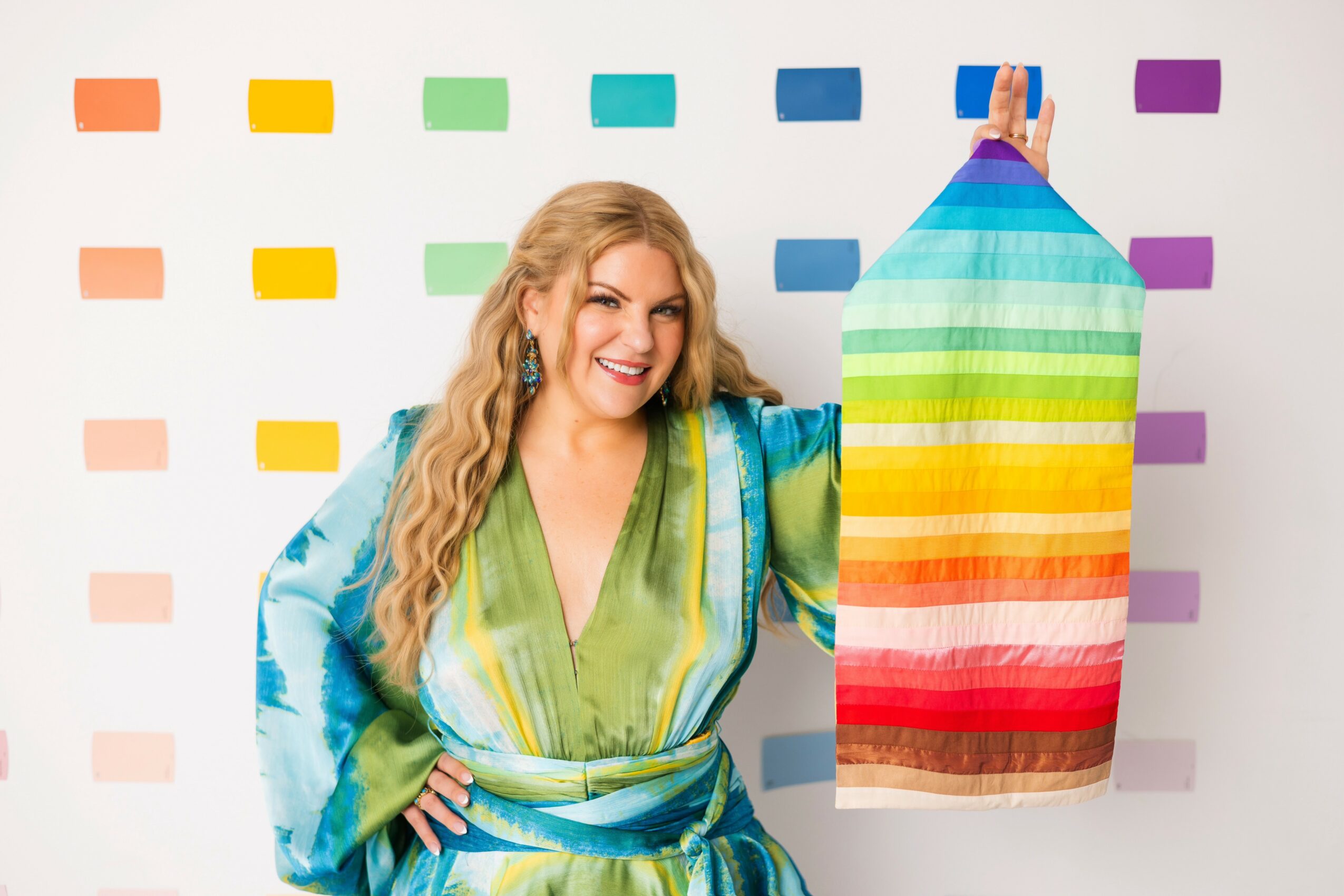 Woman with long blonde hair smiling, holding a rainbow-colored fabric, standing in front of a colorful dotted background.