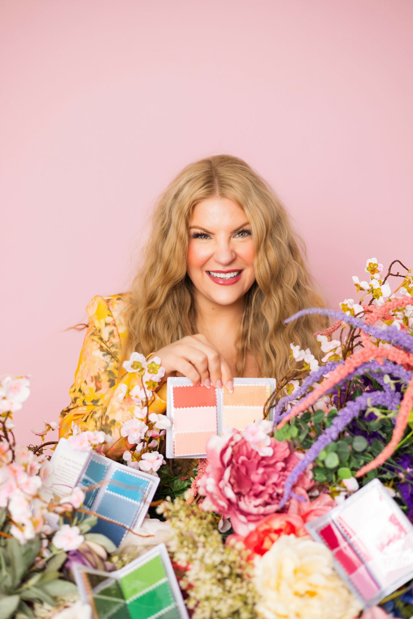 Smiling woman with long wavy hair holding a small gift box, surrounded by colorful flowers, against a pink background.