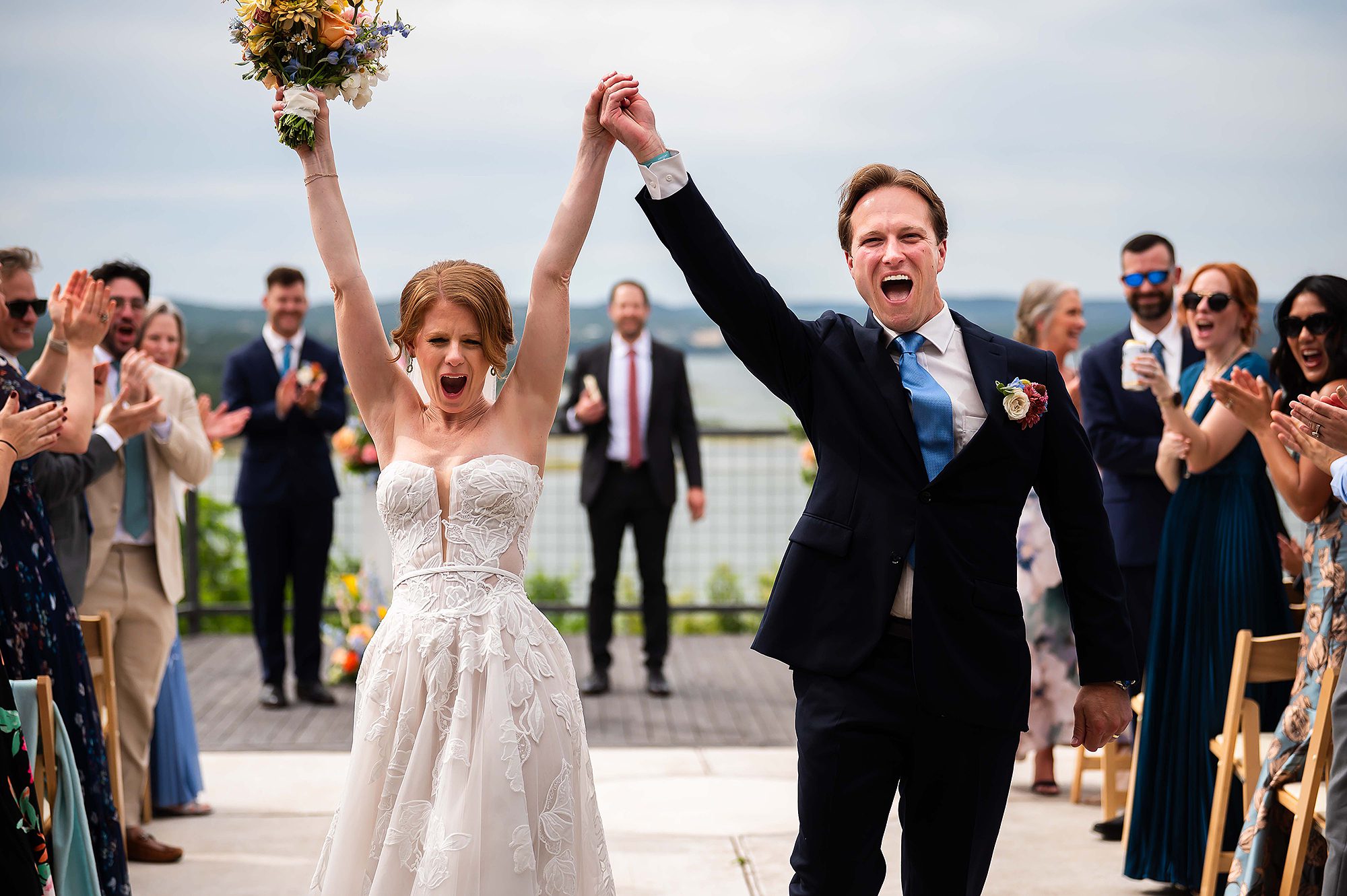 Bride and groom celebrate outdoors with raised hands, surrounded by clapping guests, on a cloudy day.