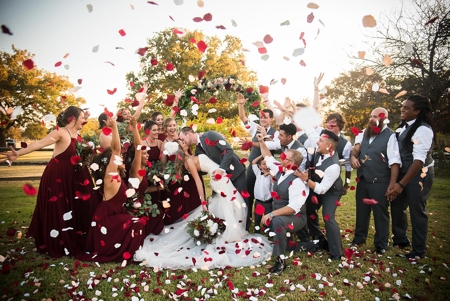Group of people celebrating outdoors with flower petals, trees, and sunlight, including a bride in a white dress and others in formal attire.