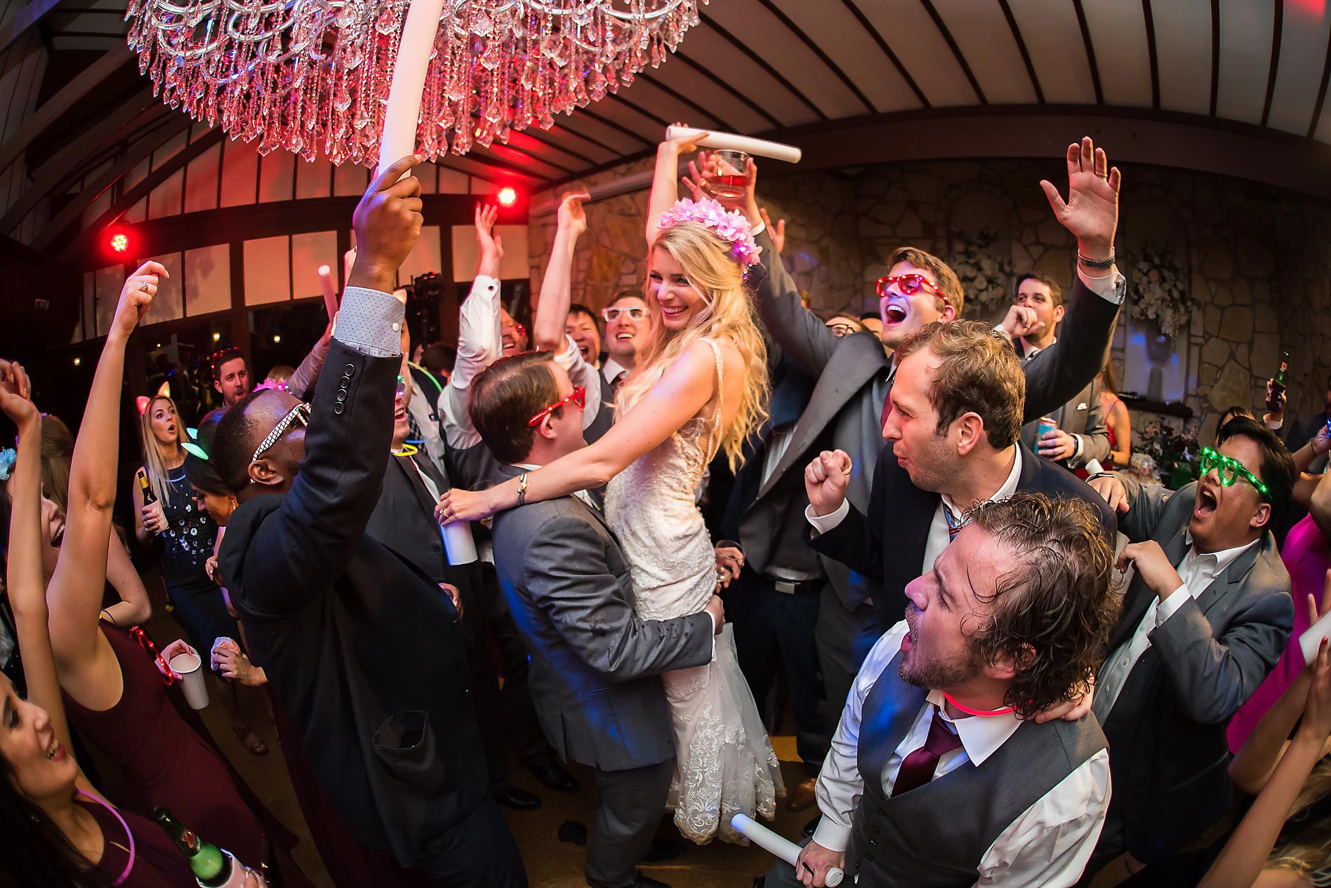 People dancing and celebrating at a party, with a woman being lifted and a chandelier overhead.