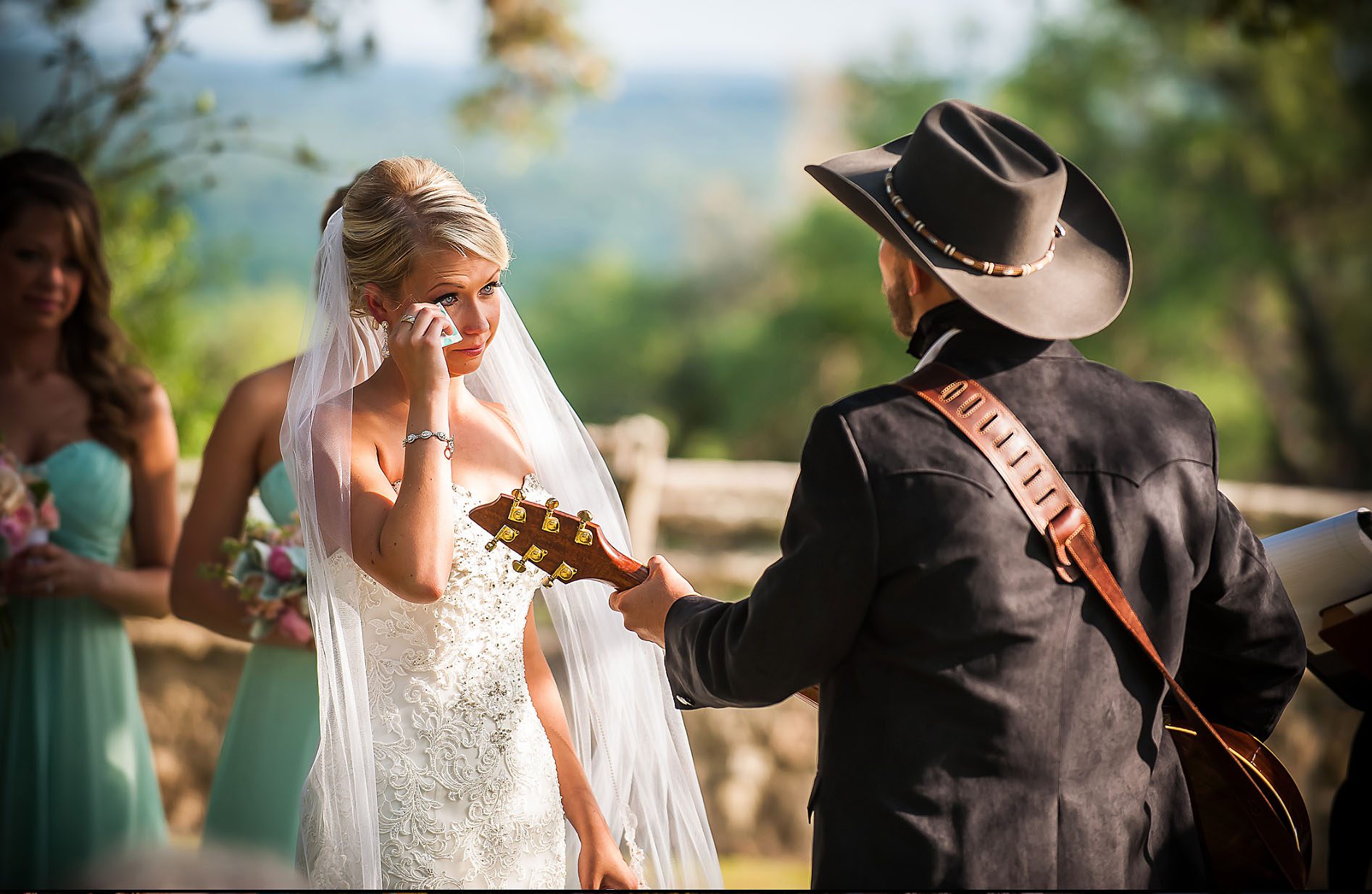 A bride in a white dress wipes tears while a man in a cowboy hat plays guitar outdoors, with a woman in a green dress nearby.