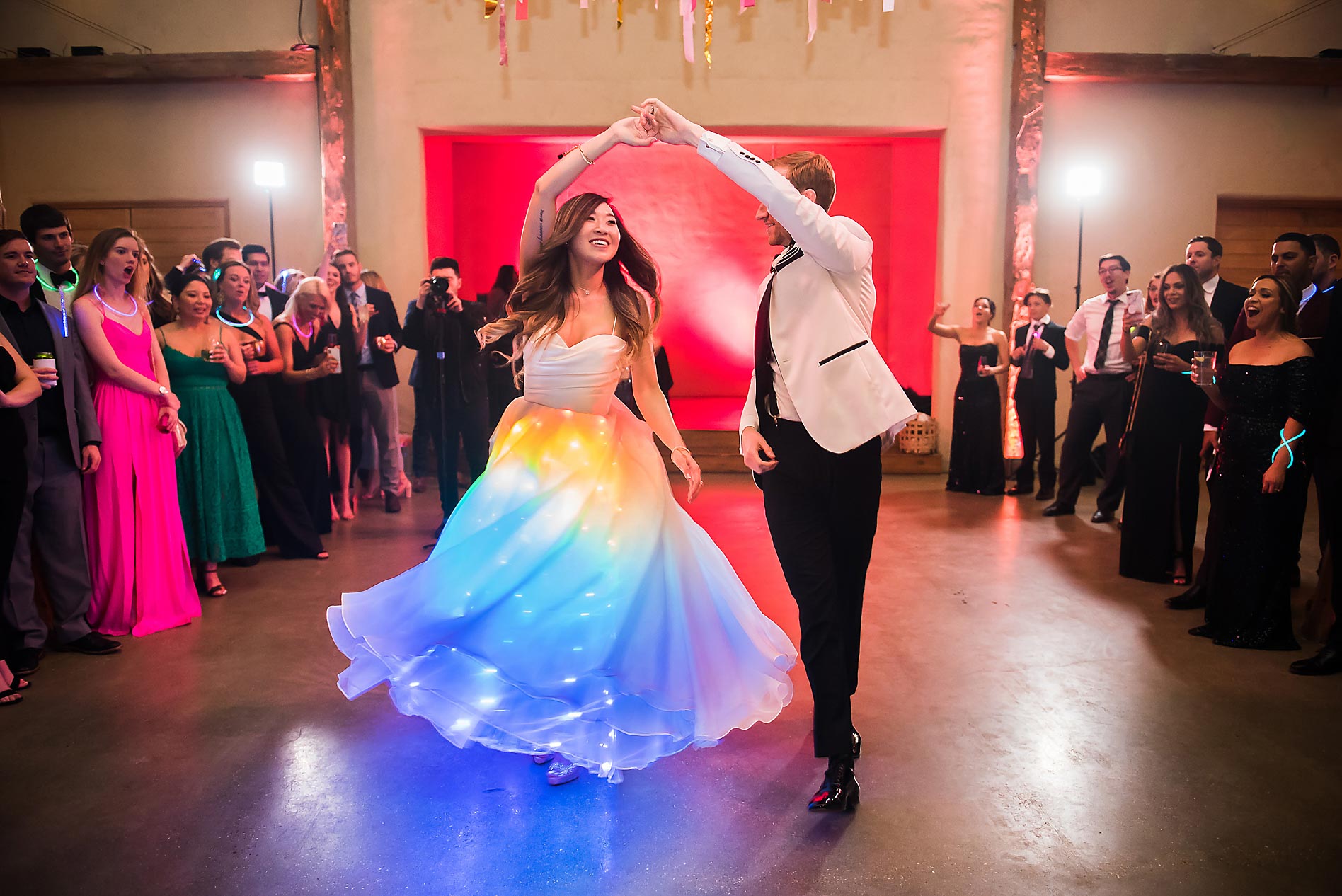 Couple dancing in center of ballroom with guests watching, woman in colorful dress and man in white jacket, decorated background.