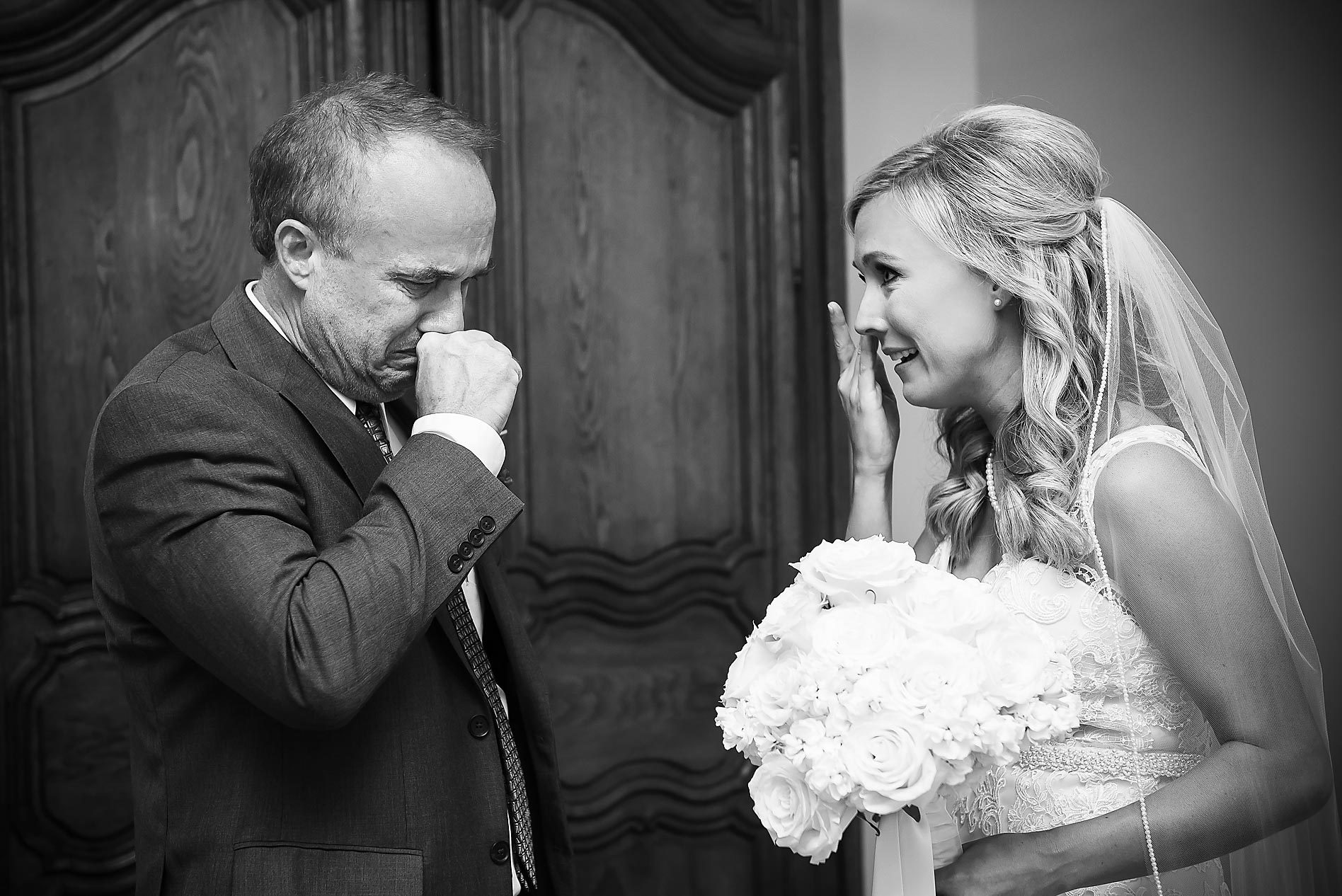 A bride holding a bouquet smiles at a man who appears emotional, in front of a wooden door, in black and white.